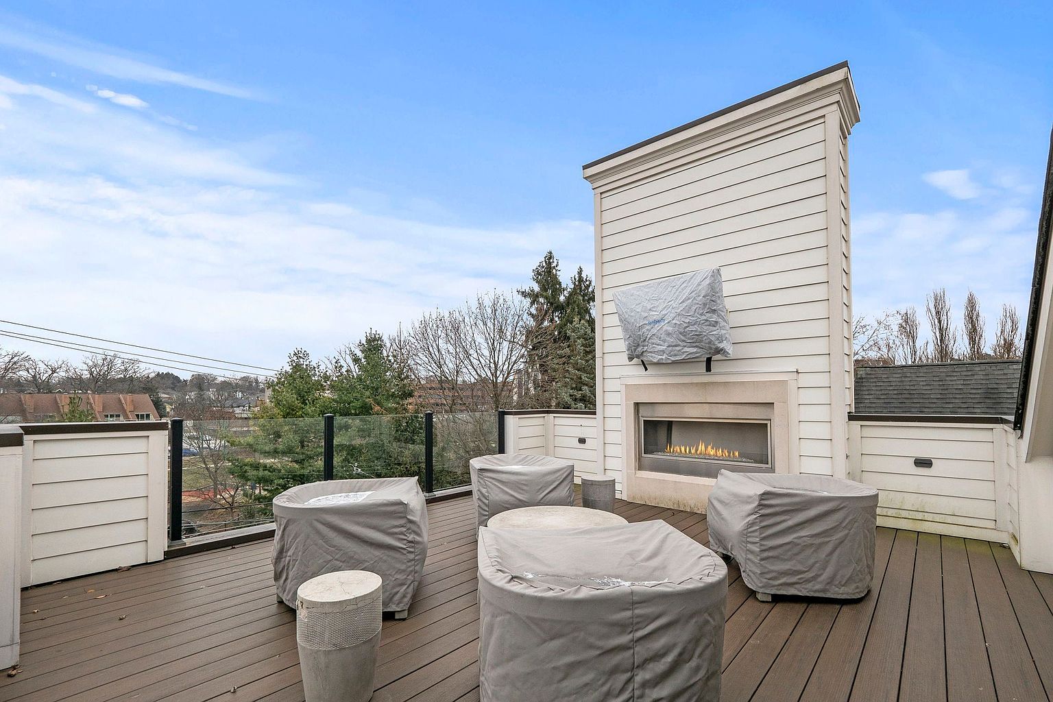 This is a view of an outdoor patio or deck area, featuring modern outdoor furniture covered with protective tarps. A sleek, built-in fireplace with a television above it serves as a focal point, adding warmth and entertainment value to the space. The deck is constructed with dark wood planks, and a glass railing provides an unobstructed view of the surrounding landscape.