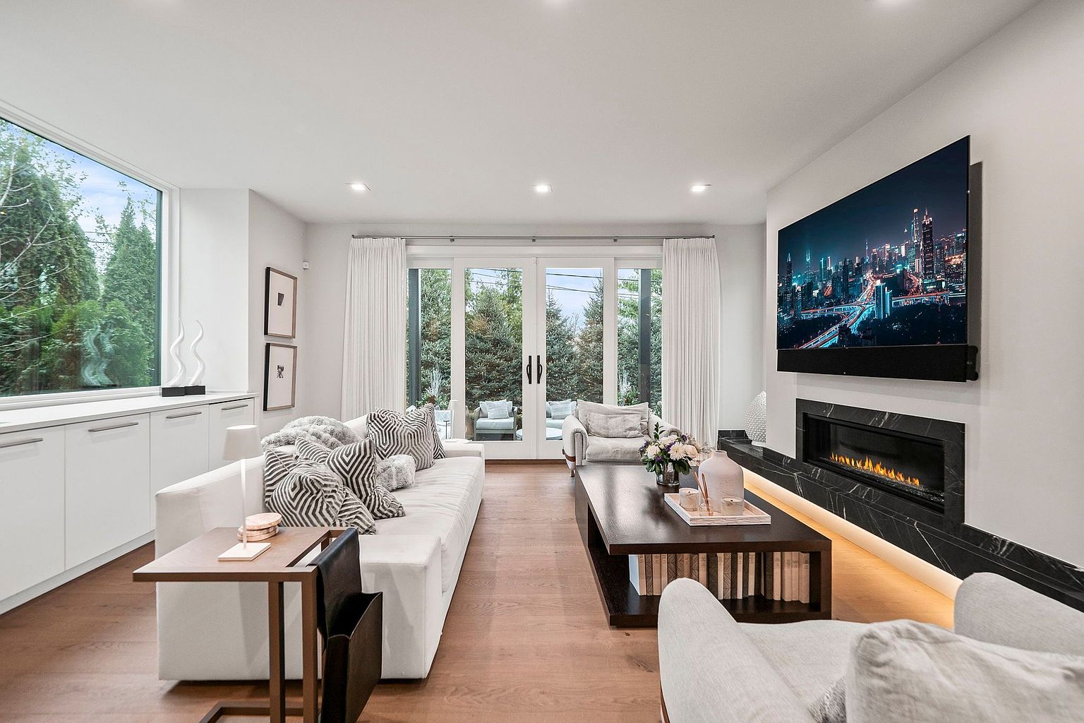 This is a bright and modern living room featuring a white sofa adorned with patterned pillows, a dark wood coffee table with books, and a sleek fireplace with a large television mounted above. The room is illuminated by natural light streaming through large windows and glass doors, which lead to an outdoor area. The hardwood floors and minimalist decor contribute to a clean and sophisticated aesthetic.