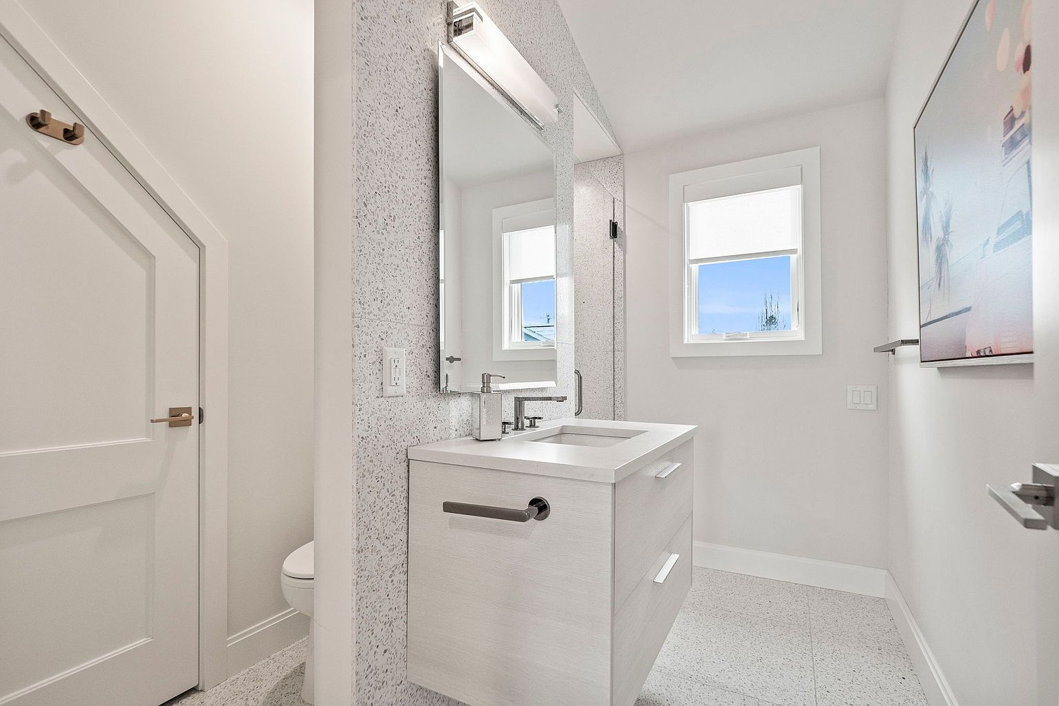This is a well-lit guest bathroom featuring a modern floating vanity with a light wood finish and a sleek countertop. A large mirror with integrated lighting hangs above the sink, and the walls are painted in a neutral tone with a textured accent wall. The floor is tiled with a speckled pattern, and a white door is visible on the left.