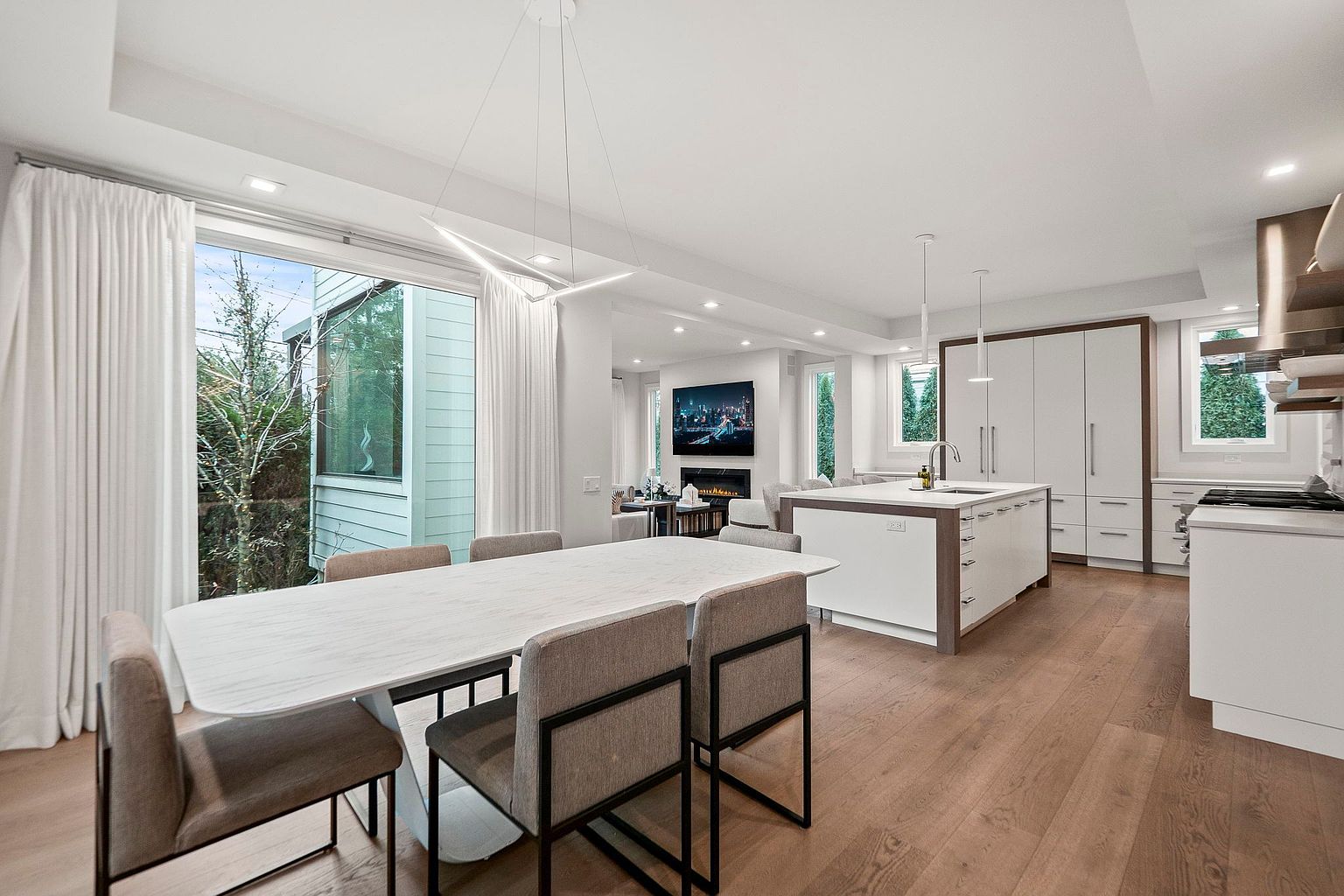 This is a bright and modern kitchen and dining area. The kitchen features white cabinetry, a large island with a sink, and stainless steel appliances. The dining area includes a white marble table with seating for six, illuminated by a modern chandelier, with a large window providing natural light and views of the outdoors.