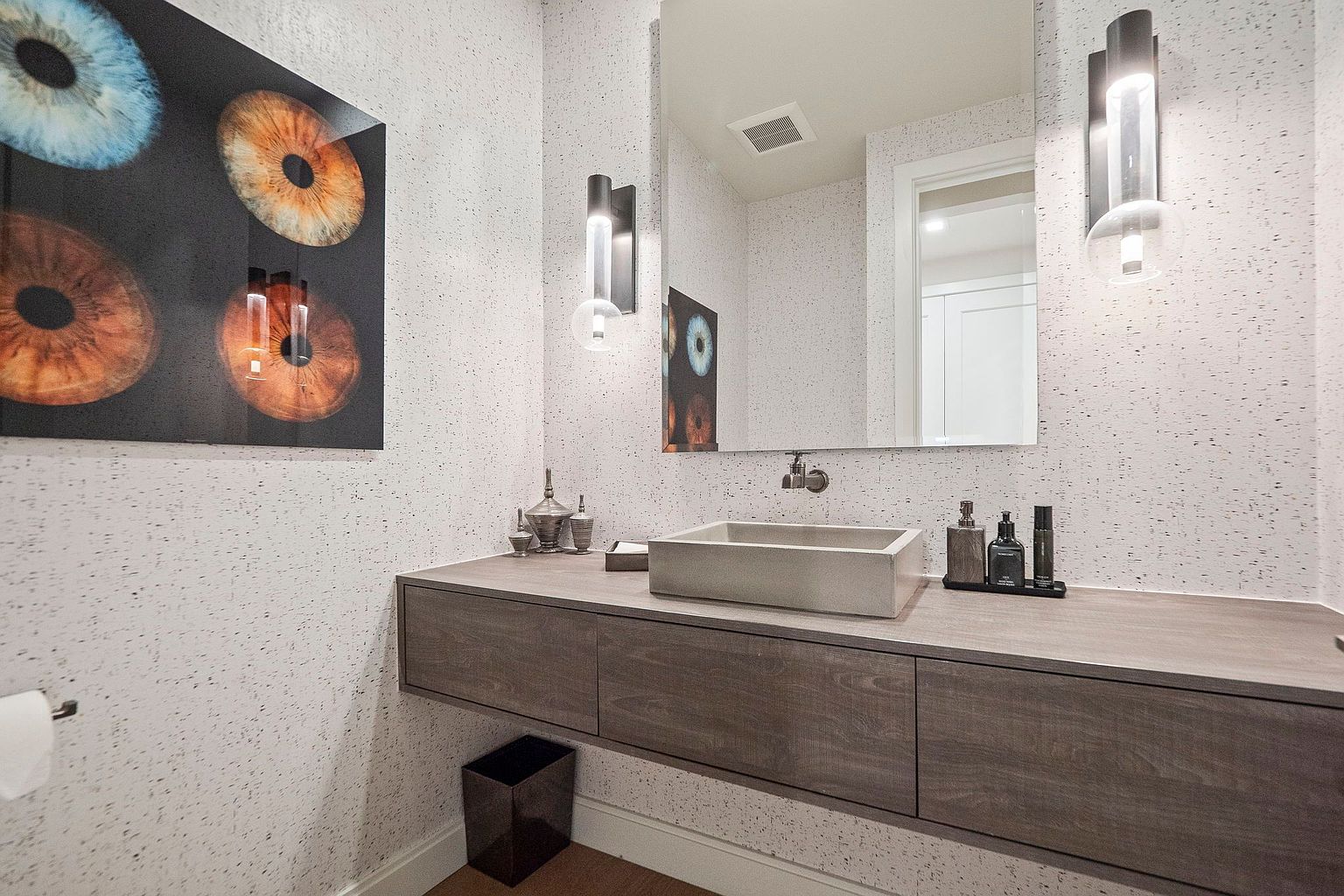 This is a modern guest bathroom featuring a floating vanity with a concrete sink and sleek, dark wood cabinetry. The walls are covered in a speckled wallpaper, and the space is illuminated by contemporary sconces flanking a large mirror. An abstract art piece adds a touch of sophistication to the room.