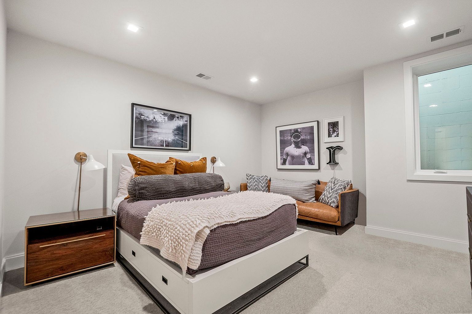 This is a modern guest bedroom featuring a bed with a white frame and gray bedding, complemented by orange pillows and a textured throw. A wooden nightstand with a lamp sits beside the bed, while a small leather couch with decorative pillows is positioned against the wall, adorned with framed artwork. The room is well-lit with recessed lighting and a window, creating a clean and inviting atmosphere.