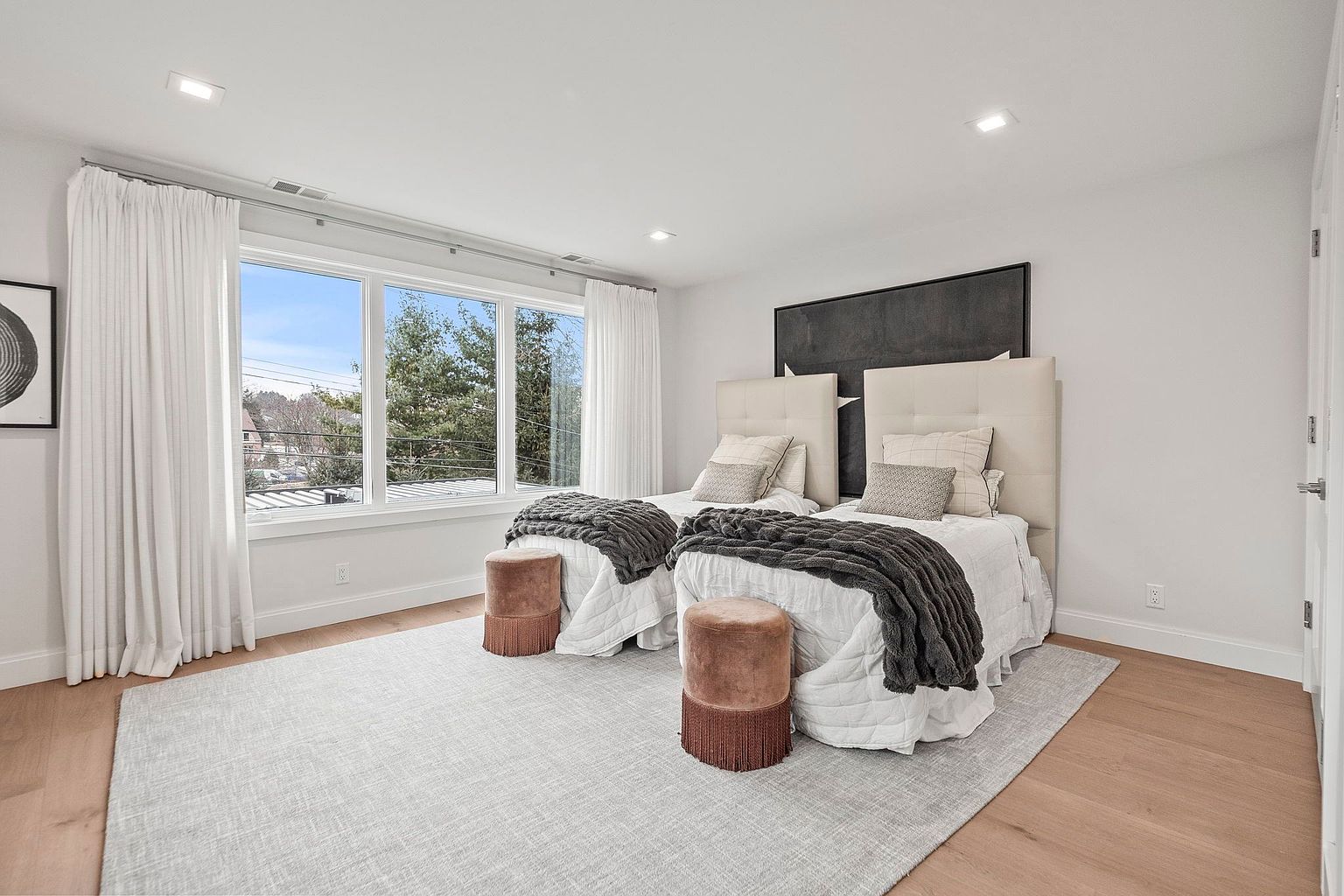 This is a bright and airy guest bedroom featuring two twin beds with upholstered headboards and neutral bedding. The room is well-lit by a large window with white curtains, and a gray rug covers the hardwood floor. Two velvet stools with fringe detail add a touch of luxury, and a large black and white artwork hangs above the beds.