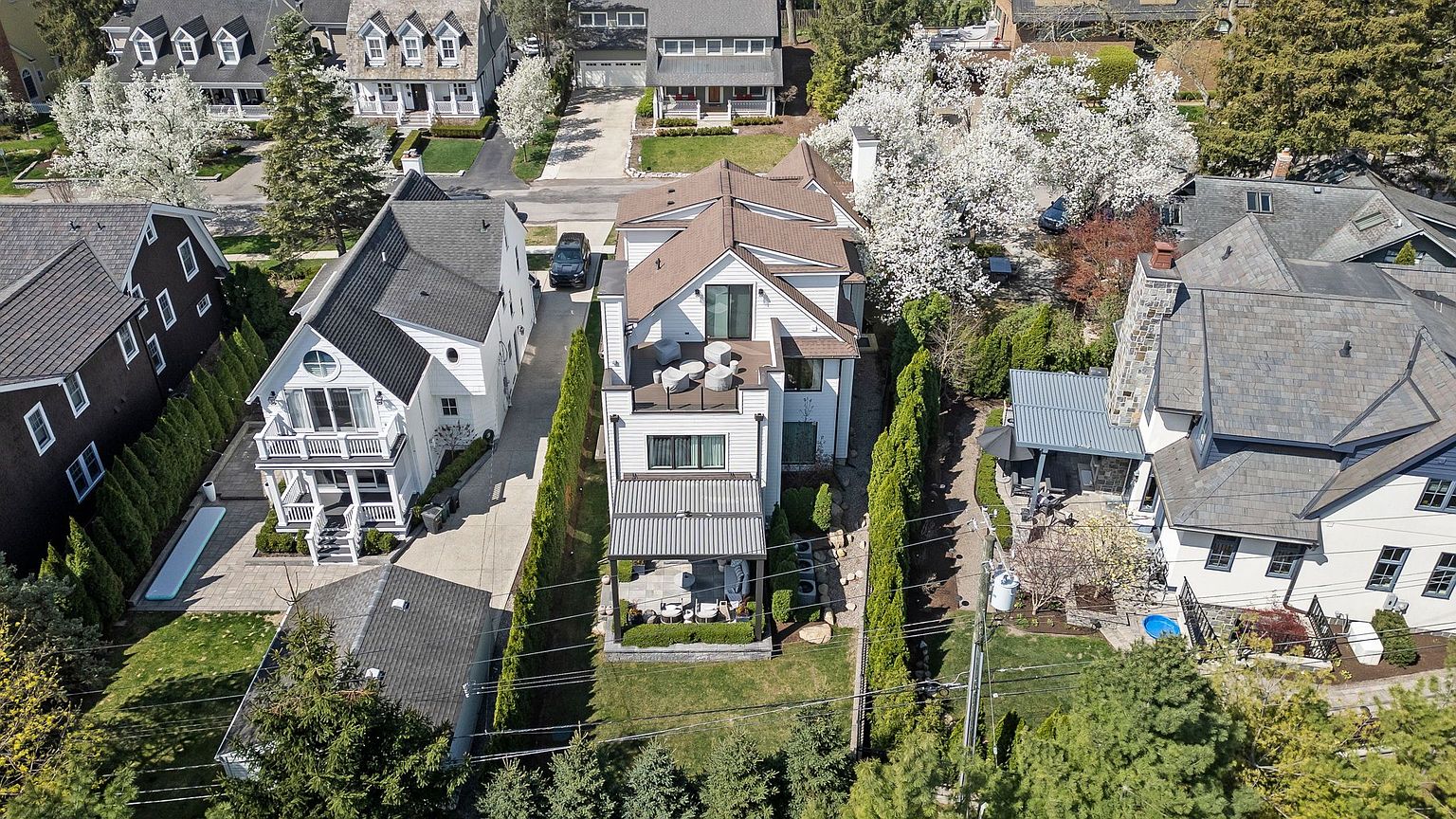 This aerial shot showcases a well-manicured property featuring a modern home with a rooftop deck and a covered patio area. The house is flanked by tall, slender trees, and the surrounding neighborhood includes a mix of architectural styles, highlighting the property's desirable location. A long driveway leads to the house, and the landscaping is meticulously maintained, adding to the overall curb appeal.