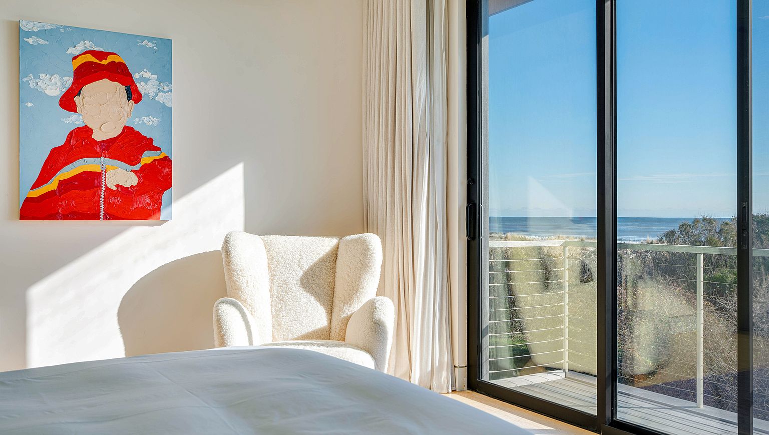 This serene bedroom features a plush, textured armchair positioned near a large sliding glass door that offers a clear view of the ocean and coastal landscape. A vibrant, abstract painting of a figure in a red jacket hangs on the wall, adding a bold pop of color to the neutral, minimalist space. The room is bathed in soft, natural light, creating a calm and inviting atmosphere perfect for relaxation.