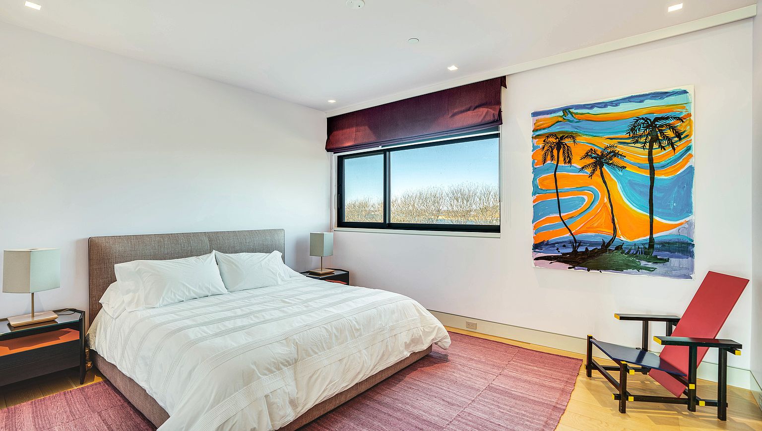 This bright and contemporary bedroom features a neutral color palette accented by a vibrant, abstract painting of palm trees on the wall. The space is furnished with a plush bed, two bedside tables with modern lamps, and a unique, geometric red lounge chair. A large window with a dark purple shade provides natural light and a view of the outdoors, while a textured purple rug adds warmth to the light wood flooring.