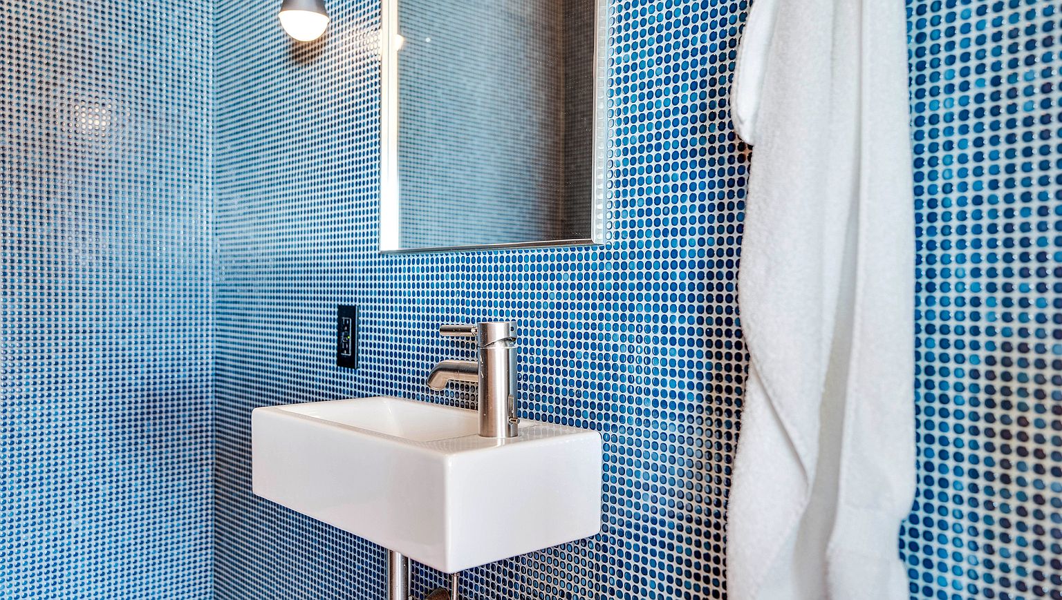 This compact bathroom features a striking wall treatment of small, circular blue mosaic tiles that create a vibrant, textured backdrop. A minimalist white wall-mounted sink is paired with a sleek, modern chrome faucet, while a simple rectangular mirror hangs above. A white towel hangs to the right, adding a soft contrast to the bold, geometric pattern of the tiled walls.