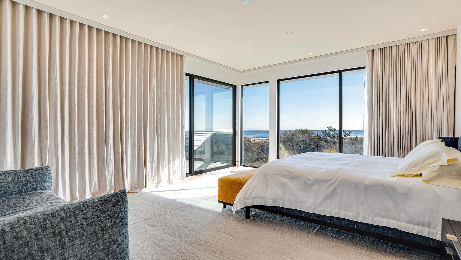 This bright and airy primary bedroom features a minimalist aesthetic with a neutral color palette and expansive floor-to-ceiling windows that offer panoramic ocean views. The room is furnished with a bed featuring a white duvet, a vibrant yellow bench at the foot, and a textured gray armchair in the foreground. Soft, light-colored curtains frame the glass walls, creating a serene and sophisticated atmosphere that emphasizes the connection to the coastal landscape.