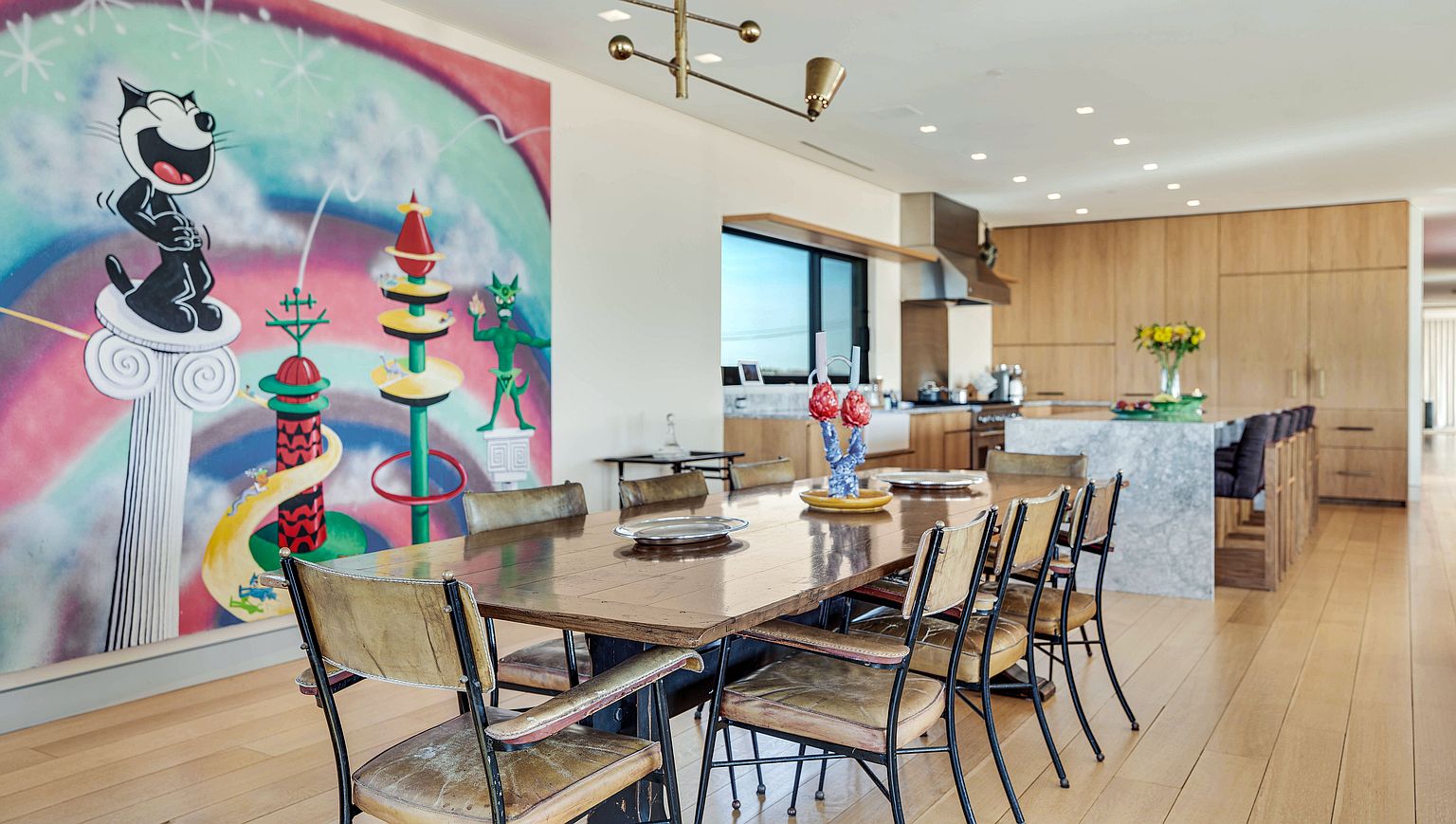 This open-concept dining area features a large, rustic wooden table surrounded by vintage-style metal chairs with worn leather upholstery. A vibrant, oversized mural of a cartoon cat dominates the wall, adding a playful and artistic focal point to the space. In the background, a modern kitchen with light wood cabinetry and a stone-topped island creates a seamless transition between dining and culinary areas.