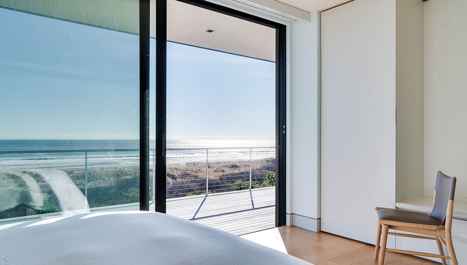 This bright bedroom features a minimalist aesthetic with light wood flooring and a sleek, modern chair positioned near a wall of white cabinetry. A large sliding glass door provides a seamless transition to an outdoor deck, framing a panoramic view of the ocean and beach. The perspective from the bed emphasizes the airy, open-concept design and the strong connection between the indoor living space and the coastal environment.