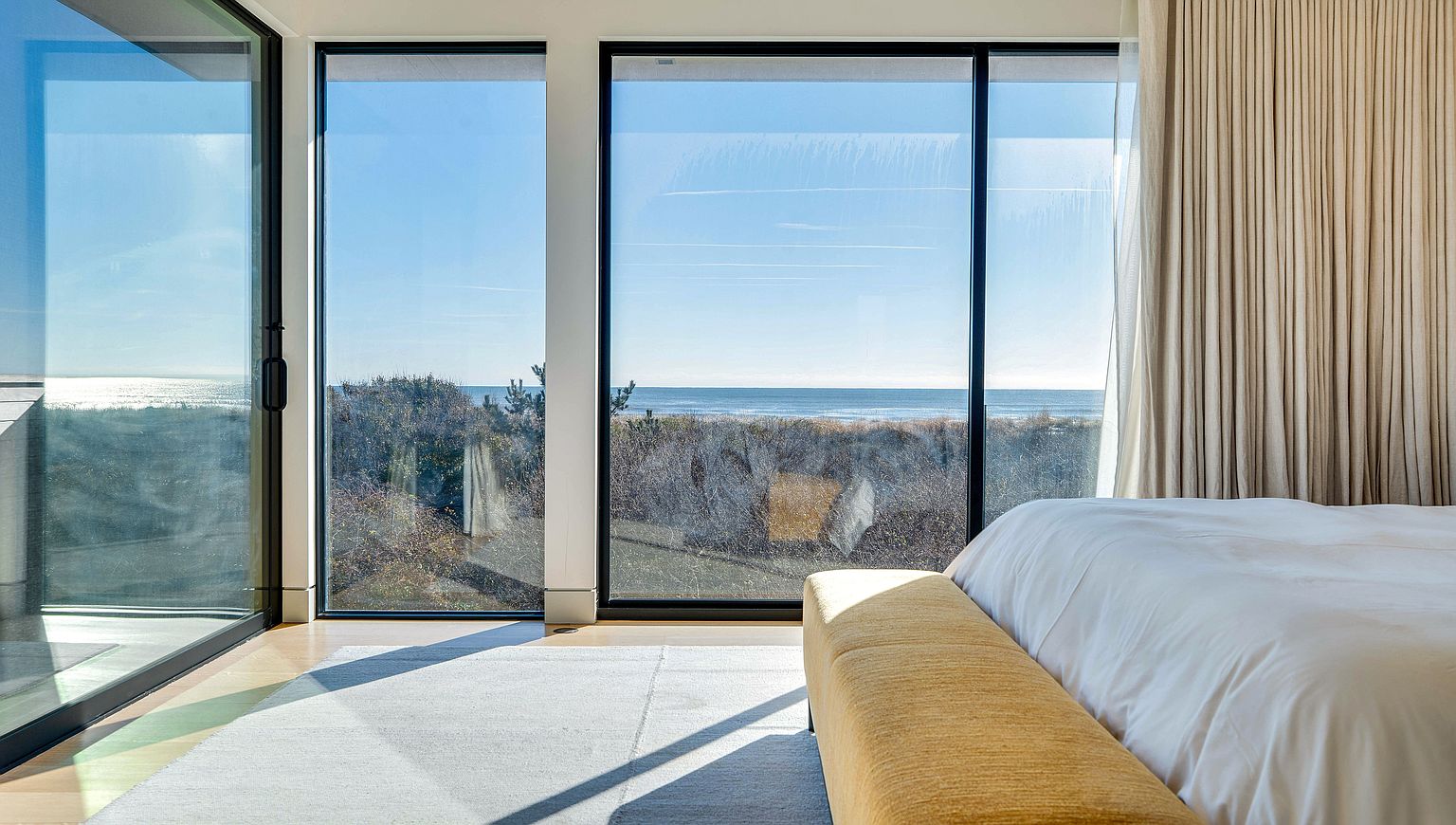 This serene primary bedroom features expansive floor-to-ceiling windows that frame a panoramic view of the ocean and coastal dunes. A plush, mustard-toned bench sits at the foot of a neatly made bed, while a light-colored area rug anchors the space. The room is bathed in natural light, creating a tranquil and airy atmosphere that emphasizes the connection between the interior living space and the natural landscape.