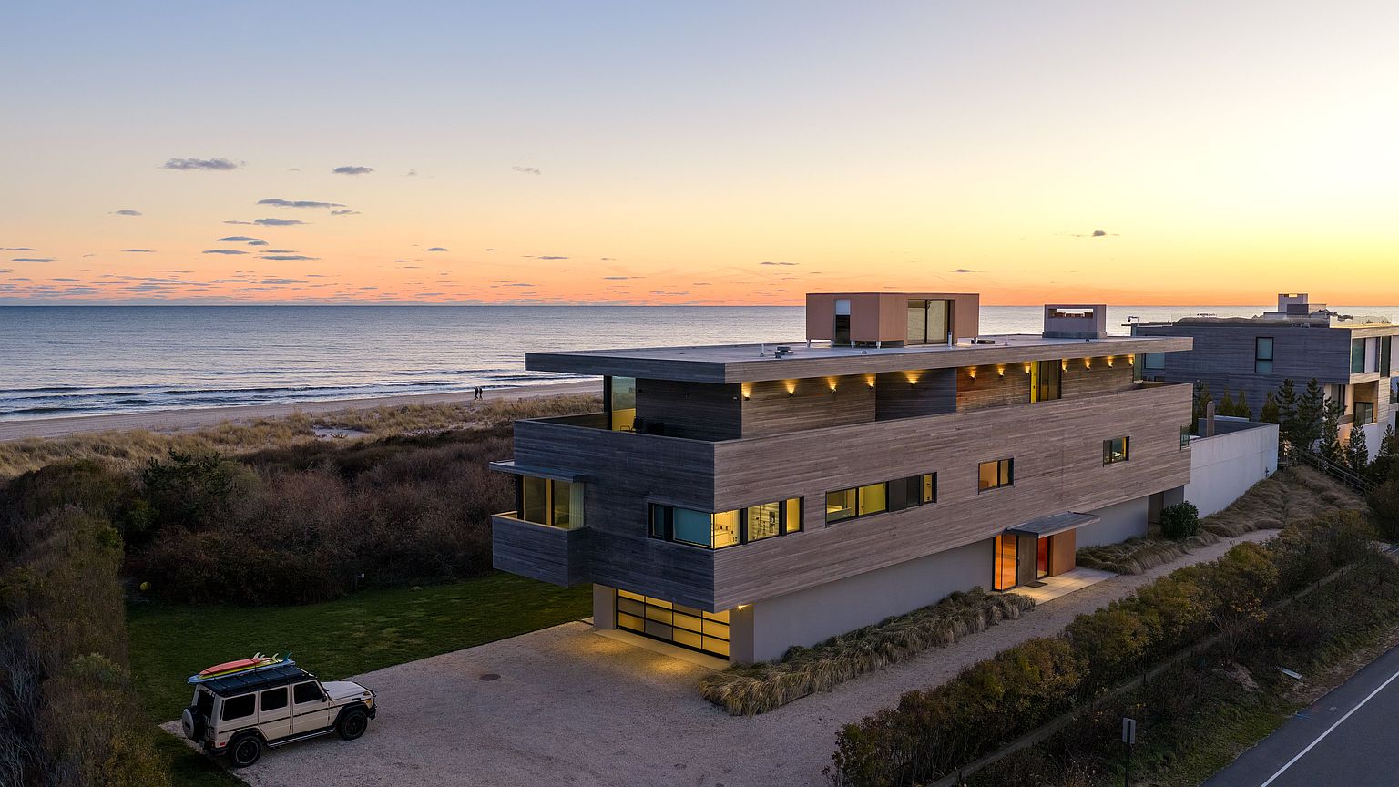 This striking aerial perspective captures a modern, multi-level coastal residence at sunset, showcasing its clean lines and wood-clad facade. The home features a prominent garage, expansive upper-level balconies, and a rooftop structure, all set against the backdrop of a serene beach and ocean horizon. A light-colored SUV with surfboards on the roof is parked on the gravel driveway, emphasizing the property's proximity to the water and its lifestyle appeal.