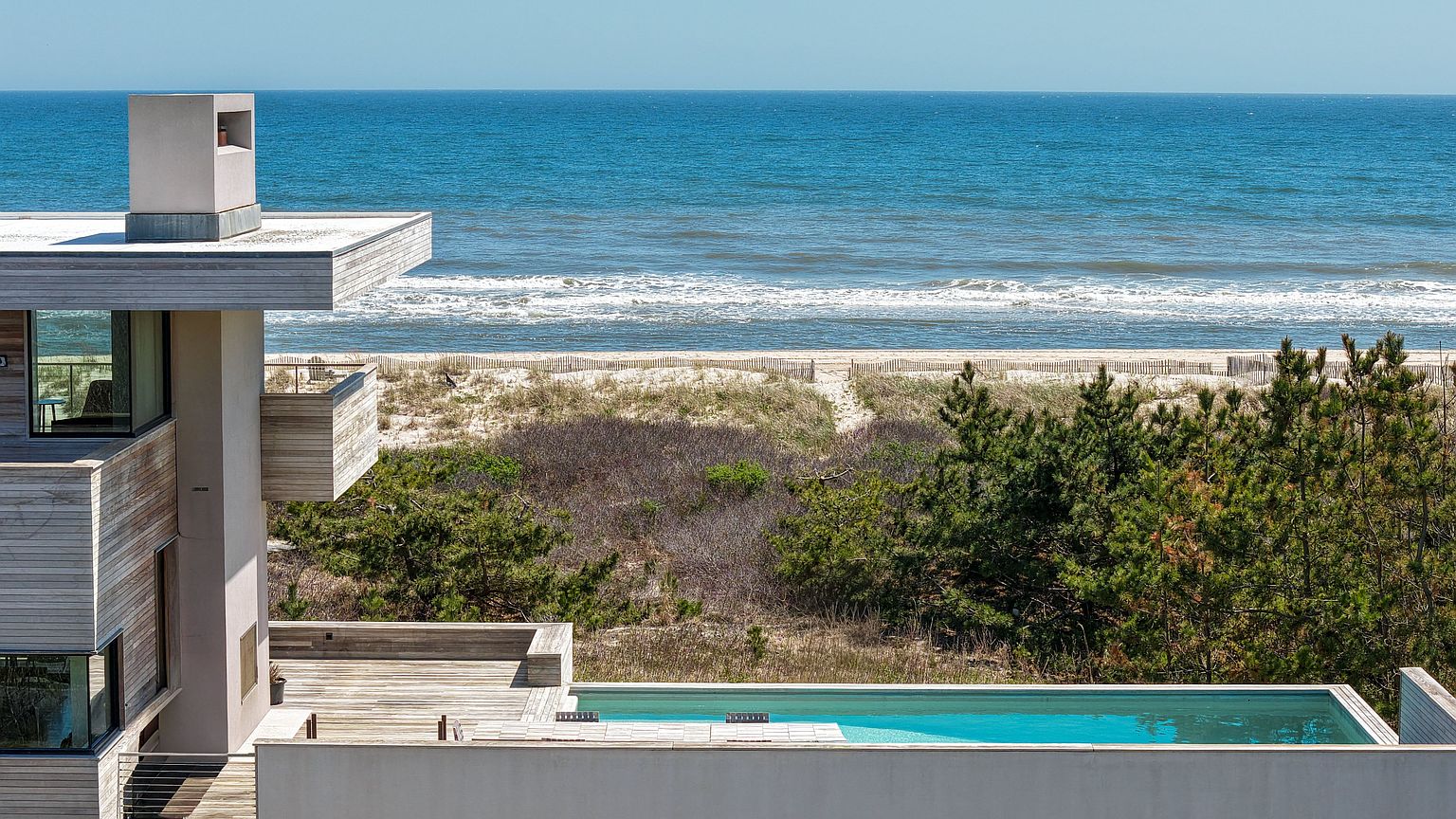 This elevated perspective captures a modern, multi-level residence featuring horizontal wood siding and a prominent chimney structure. A sleek, rectangular swimming pool sits on a terrace overlooking a lush dune landscape and the expansive ocean beyond. The composition emphasizes the seamless integration of contemporary architecture with the natural coastal environment.