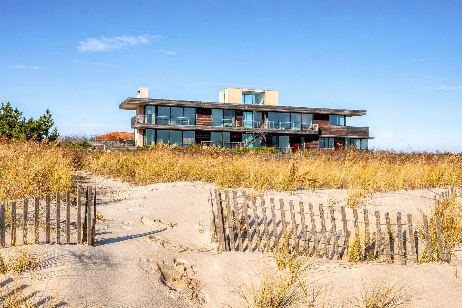 This contemporary coastal residence features a multi-level design with extensive glass walls and dark wood siding, set against a backdrop of dunes and a clear blue sky. A weathered wooden fence frames a sandy path leading toward the home, emphasizing its secluded beachside location. The architecture highlights clean horizontal lines and expansive balconies that offer panoramic views of the surrounding landscape.