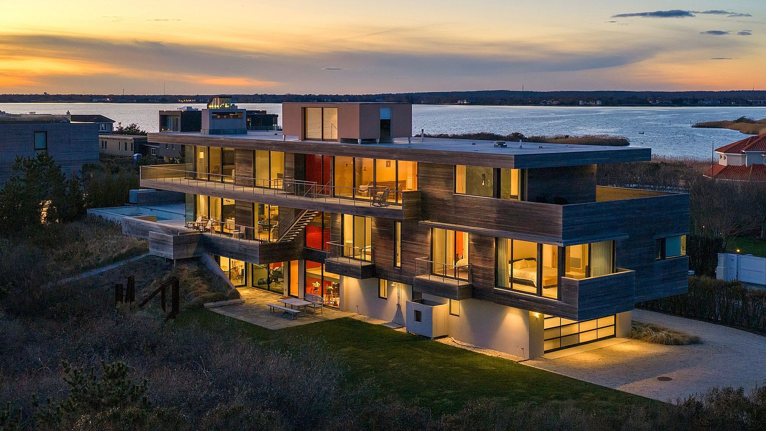 This striking modern residence is captured from an elevated aerial perspective at sunset, showcasing its multi-level architectural design and extensive use of wood siding. The home features expansive glass walls, multiple balconies, and a seamless integration with the surrounding coastal landscape, including a view of the nearby water. The warm glow from the interior lighting contrasts beautifully with the twilight sky, highlighting the property's sophisticated and contemporary aesthetic.