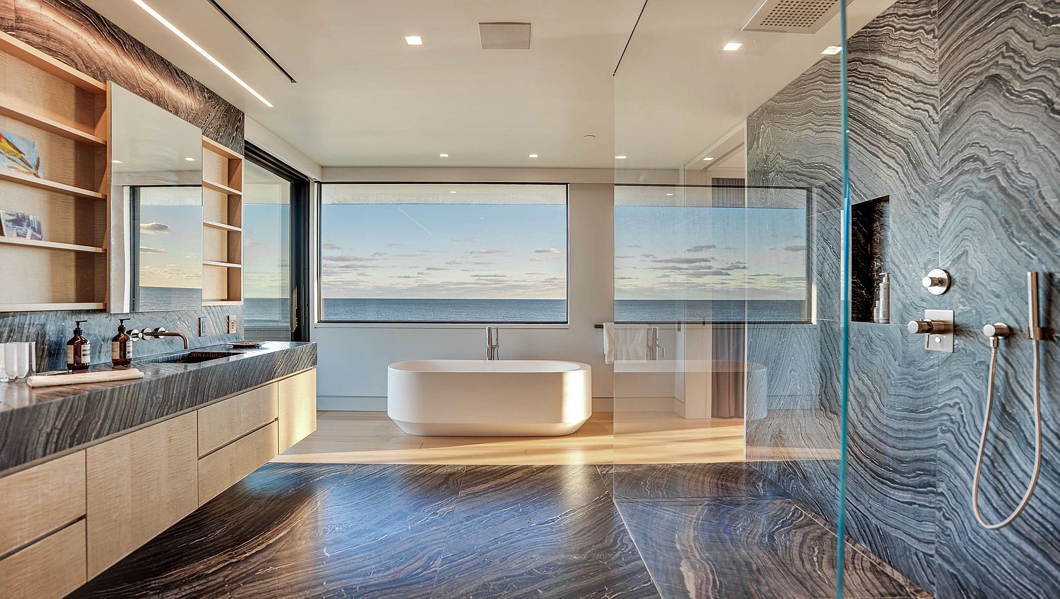 This luxurious primary bathroom features a striking, dark-veined stone aesthetic that extends across the vanity and shower walls. A freestanding white soaking tub is positioned centrally in front of a large window, offering expansive ocean views. The space is complemented by light wood cabinetry, open shelving, and a glass-enclosed shower, creating a sophisticated and serene atmosphere.
