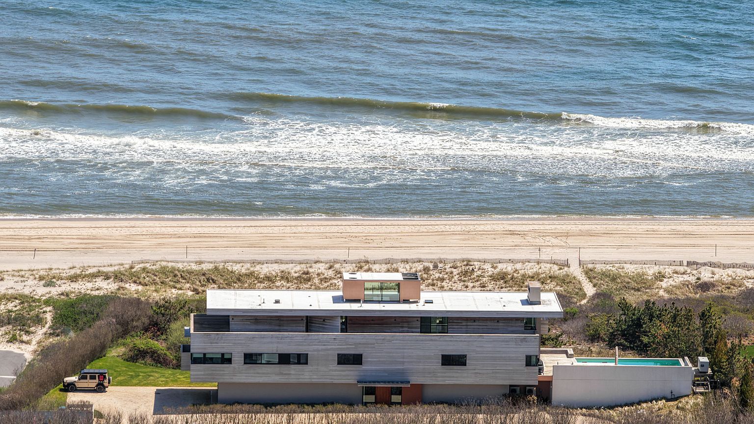 This aerial perspective captures a modern, multi-level residence situated directly on a sandy beach. The home features a sleek, horizontal design with light-toned wood siding, a flat roof with a central glass-enclosed structure, and a private swimming pool on the side. A light-colored SUV is parked on the gravel driveway, while the vast ocean waves roll onto the shore in the background.