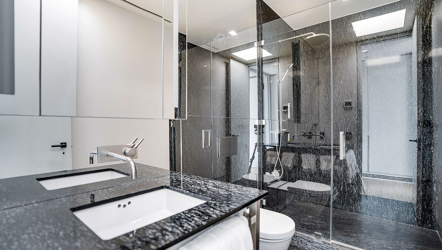 This modern bathroom features a sleek vanity with a dark, speckled stone countertop and dual white undermount sinks. A glass-enclosed shower area with dark stone walls and a rainfall showerhead creates a sophisticated, spa-like atmosphere. The space is illuminated by recessed lighting and a large mirror, reflecting the clean, contemporary design.