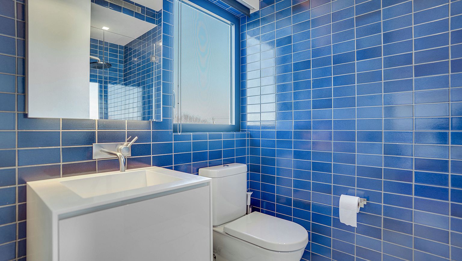 This compact bathroom features striking, vibrant blue rectangular wall tiles that create a bold, modern aesthetic. The space includes a minimalist white vanity with a sleek faucet, a wall-mounted mirror, and a standard white toilet. A window with a roller blind allows natural light to brighten the room, while the clean lines and high-contrast color palette provide a contemporary feel.