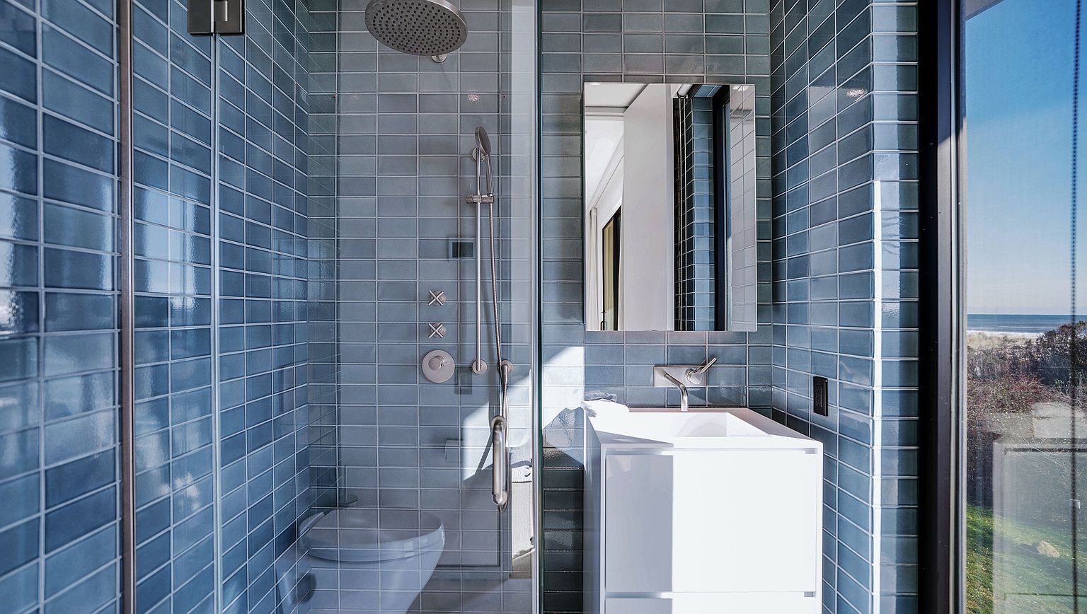 This compact, modern bathroom features striking blue subway-style wall tiles that create a cohesive and vibrant aesthetic. The space includes a glass-enclosed shower area with a rainfall showerhead and a sleek, white vanity with a wall-mounted faucet. A large window to the right provides natural light and a view of the outdoors, enhancing the clean, contemporary feel of the room.