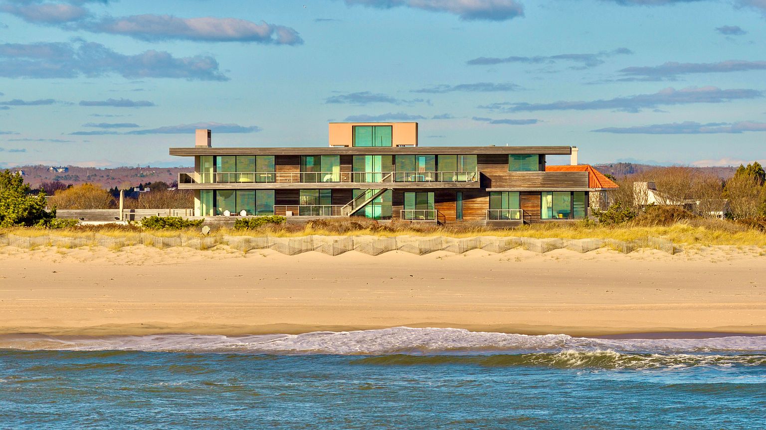 This modern, two-story beachfront residence features a horizontal wood-clad facade and extensive glass glazing that offers panoramic ocean views. The home is situated directly behind a sandy dune, with a wooden walkway and tiered balconies providing seamless indoor-outdoor living. The architectural style is minimalist and contemporary, emphasizing clean lines and a strong connection to the coastal landscape.