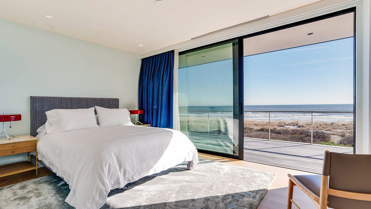 This bright primary bedroom features a minimalist aesthetic with a plush bed centered against a light-colored wall. A large sliding glass door provides an expansive, unobstructed view of the ocean and the adjacent wooden deck. The room is accented by deep blue curtains and modern red bedside lamps, creating a serene and sophisticated atmosphere.