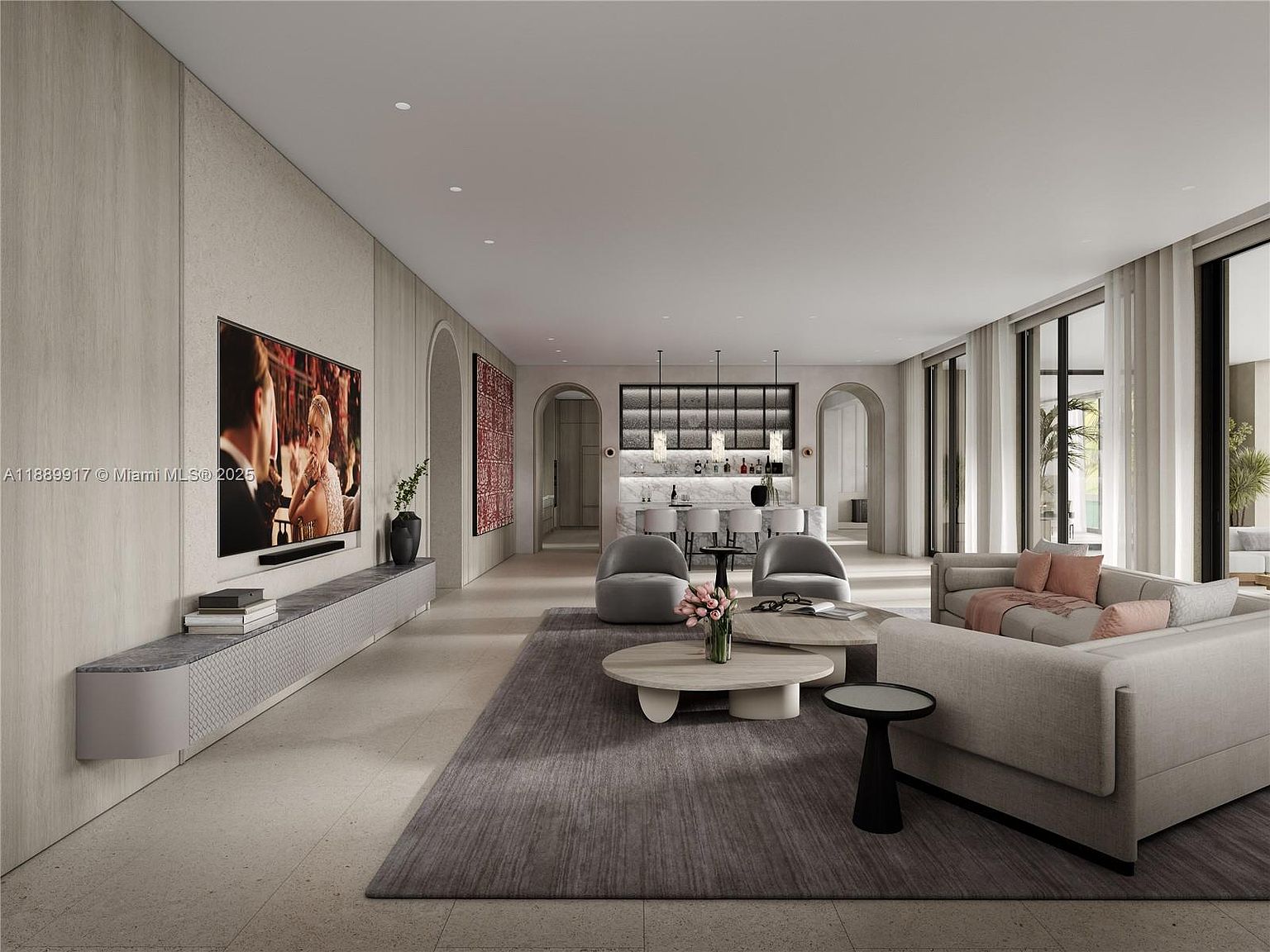 This is a spacious and modern living room featuring a large sectional sofa, a round coffee table, and a flat-screen TV mounted on the wall. The room has a neutral color palette with gray and beige tones, complemented by natural light streaming through large windows. The open floor plan extends to a bar area, creating a seamless and inviting living space.