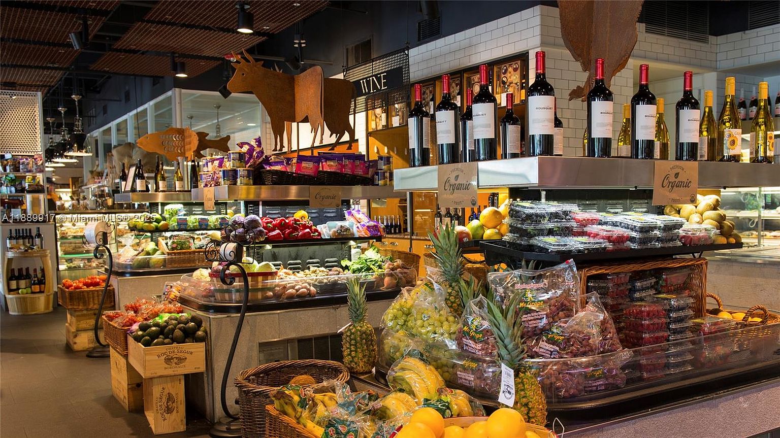 This image showcases the interior of a high-end grocery store, focusing on the produce and wine sections. The display features a variety of fresh fruits, vegetables, and bottled wines, arranged attractively on tiered shelves and in baskets. The lighting is bright and inviting, highlighting the quality and freshness of the products, creating an upscale shopping experience.