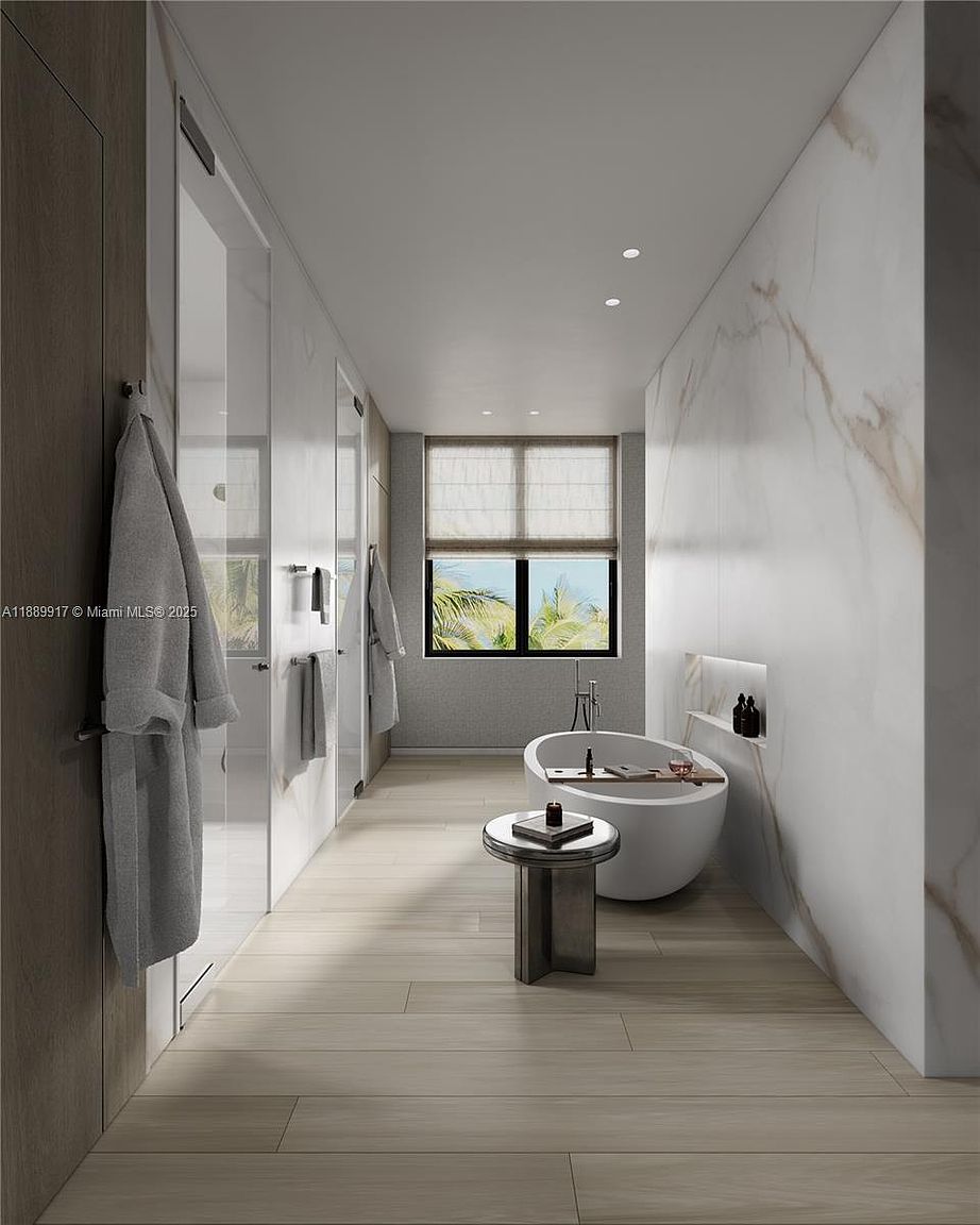 This is a modern primary bathroom featuring a freestanding bathtub with a small side table, a large window with a view of palm trees, and a walk-in shower. The walls are clad in marble, and the floor is tiled with light wood-look planks. The perspective is a long shot, emphasizing the length of the bathroom and the clean, minimalist design.