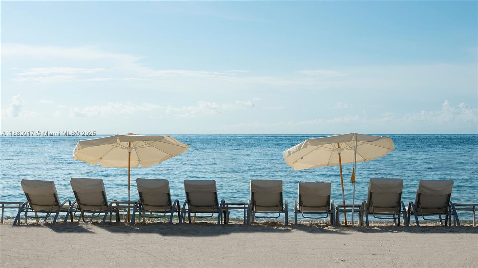 This image showcases a serene beachfront patio or deck area, featuring multiple lounge chairs arranged in a row, each set complemented by a small table. Two large white umbrellas provide shade, enhancing the comfort and appeal of the space. The backdrop is a tranquil ocean view under a clear blue sky, creating an inviting and relaxing atmosphere.