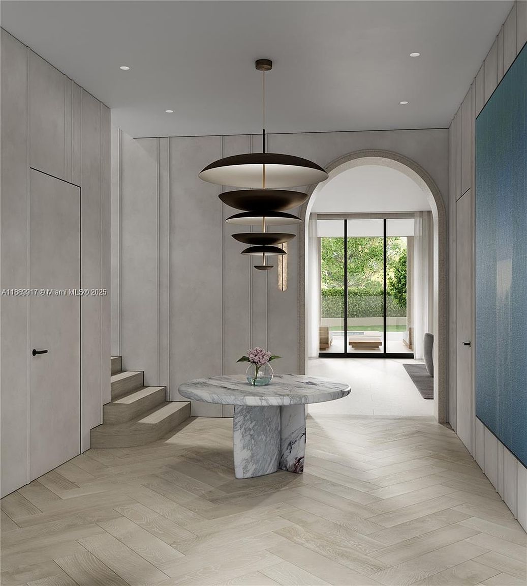 This interior shot showcases a modern hallway with herringbone wood flooring, a marble round table with a floral arrangement, and a unique tiered pendant light fixture. The hallway features clean lines, neutral tones, and an arched doorway leading to an outdoor view, creating a sophisticated and inviting atmosphere. A staircase is partially visible, suggesting multiple levels in the home.