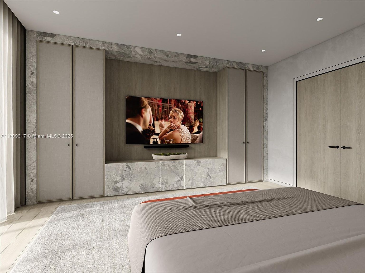 This is a primary bedroom featuring a large television mounted on a wood-paneled wall, flanked by built-in wardrobes with light gray doors and marble accents. A low marble cabinet sits beneath the TV, and the bed is partially visible in the foreground, suggesting a spacious and modern design. The room has a minimalist aesthetic with neutral tones and clean lines.