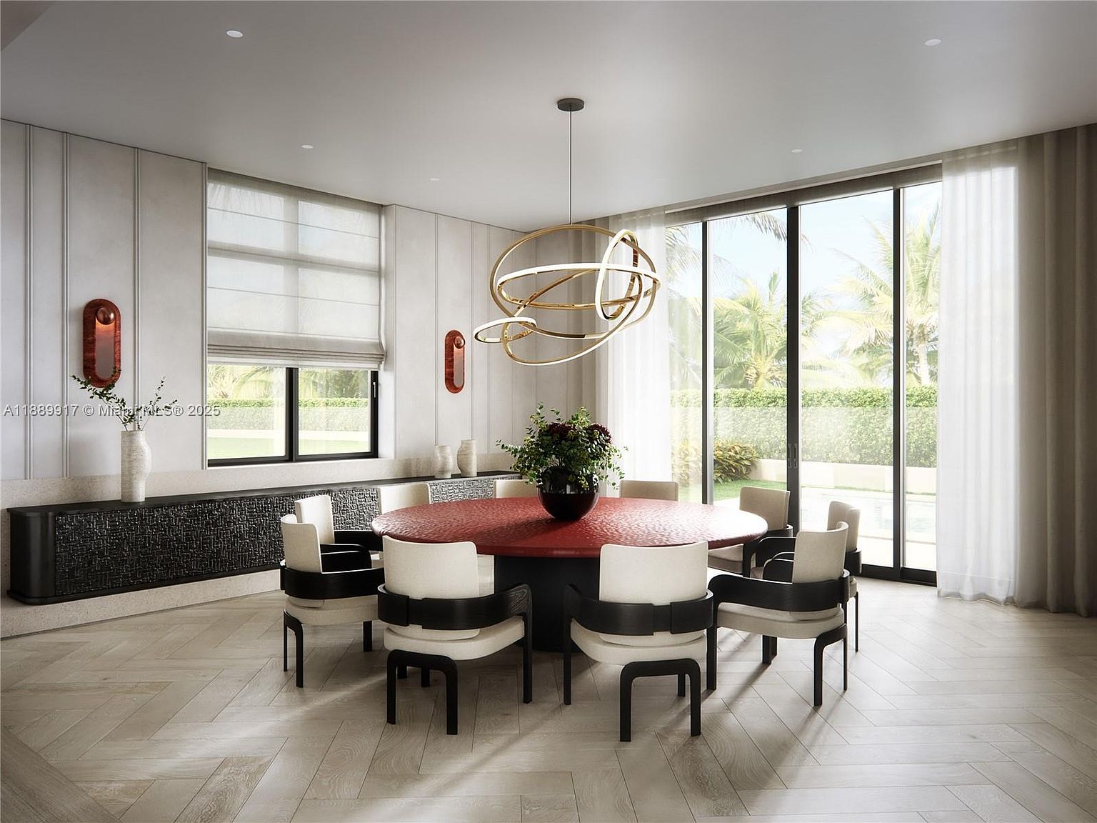 This is a dining room featuring a large, round red table surrounded by eight chairs with black and white accents. A modern, multi-ring chandelier hangs above the table, and large windows offer a view of the exterior greenery. The room is decorated in a contemporary style with neutral tones and sleek lines, creating an elegant and inviting atmosphere.