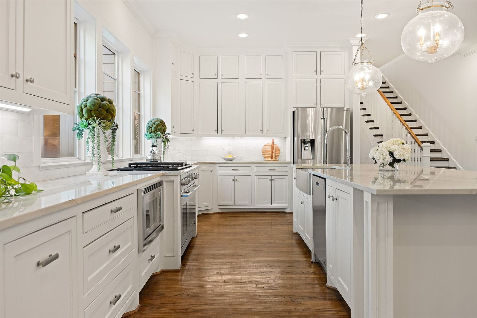 This is a bright and spacious kitchen featuring white cabinetry, stainless steel appliances, and a large center island with a light-colored countertop. The hardwood floors add warmth to the space, while the pendant lighting and recessed lights create a welcoming atmosphere. A staircase is visible in the background, suggesting an open floor plan.