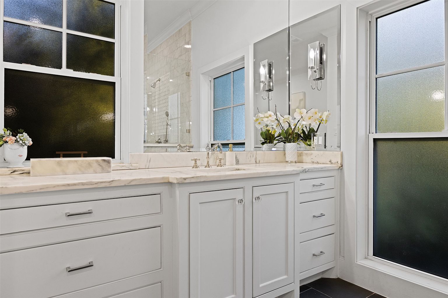 This is a well-lit primary bathroom featuring a double vanity with a marble countertop and white cabinetry. The vanity has decorative crystal knobs and pulls, and the mirrors are flanked by sconces. The room has a clean and luxurious feel, with a window providing natural light and a glimpse of the shower area.