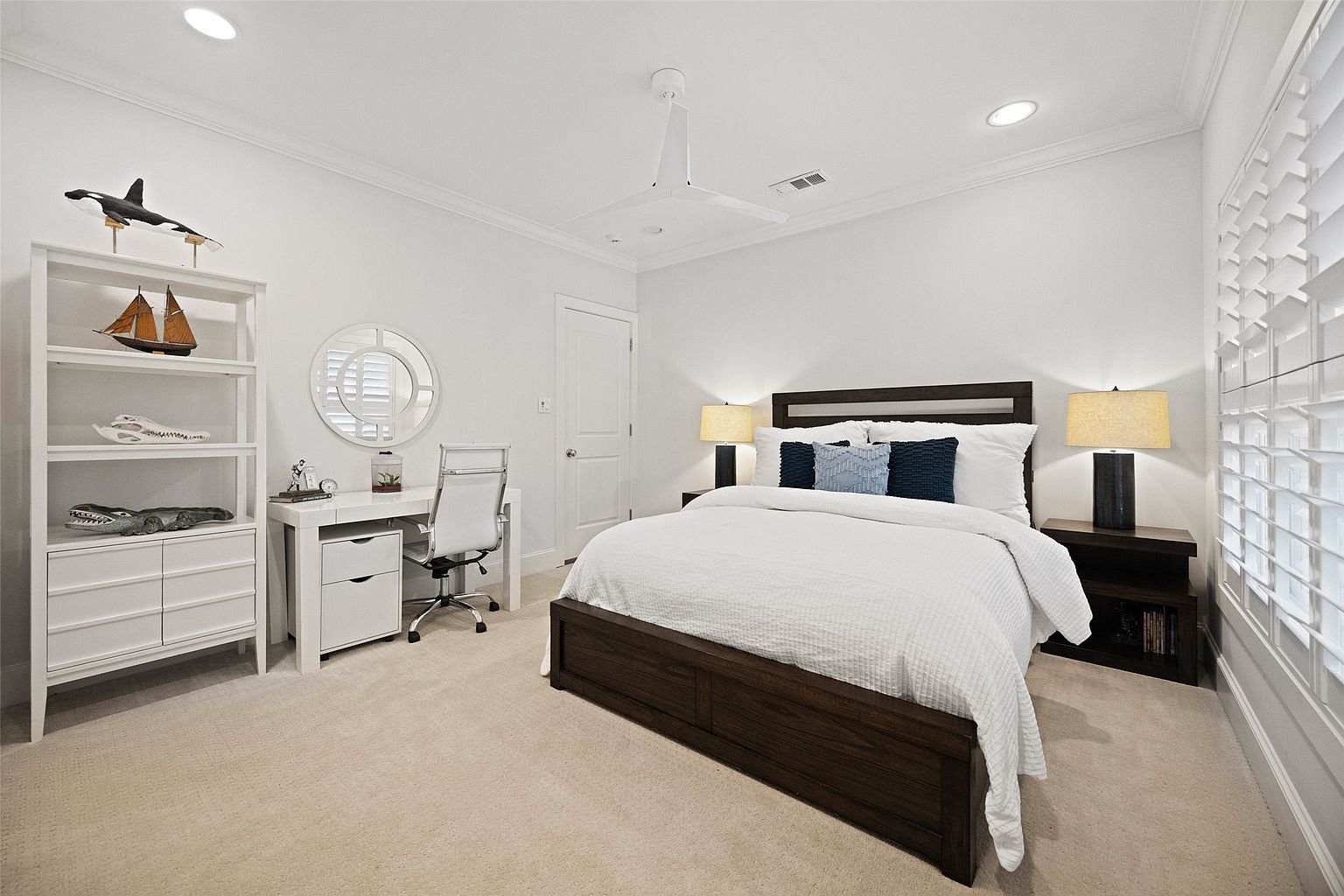 This is a well-lit bedroom featuring a dark wood bed frame with white bedding and blue accent pillows. A white desk and chair are positioned near a round mirror and a white shelving unit displaying decorative items. The room has a clean and modern aesthetic with white walls and neutral carpet.