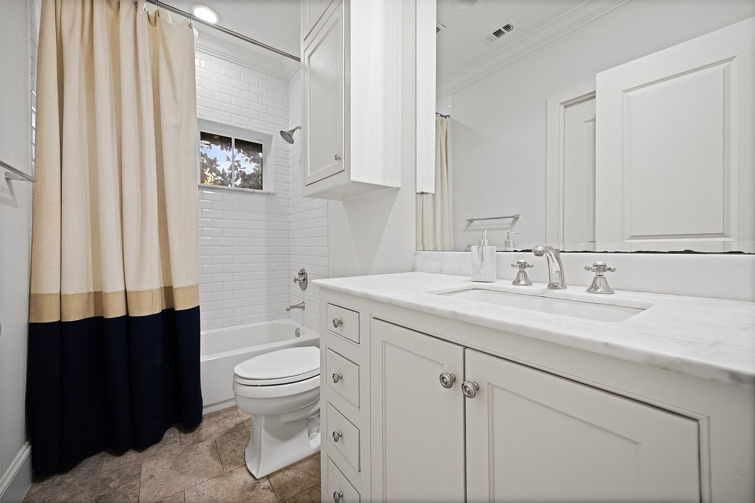 This is a well-lit bathroom featuring a white vanity with a marble countertop, a sink, and chrome fixtures. A toilet is positioned next to the bathtub, which has a shower and a decorative shower curtain. The walls are tiled in a classic subway pattern, and the flooring appears to be a neutral tile.