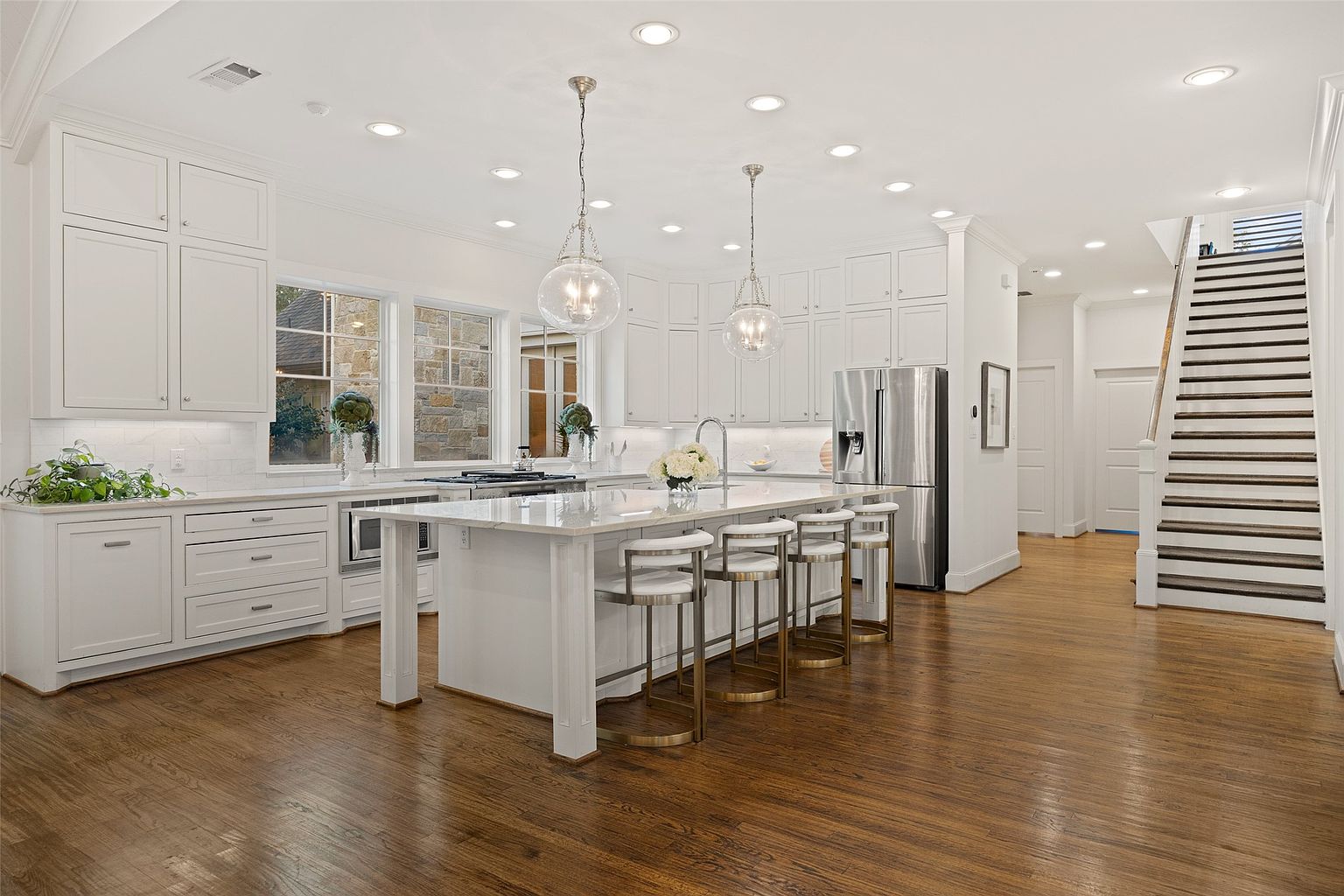 This is a bright and spacious kitchen featuring white cabinetry, stainless steel appliances, and a large center island with seating. The kitchen is well-lit with recessed lighting and pendant lights over the island, and it has hardwood floors. The perspective is from the entrance of the kitchen, showcasing the island, appliances, and staircase.
