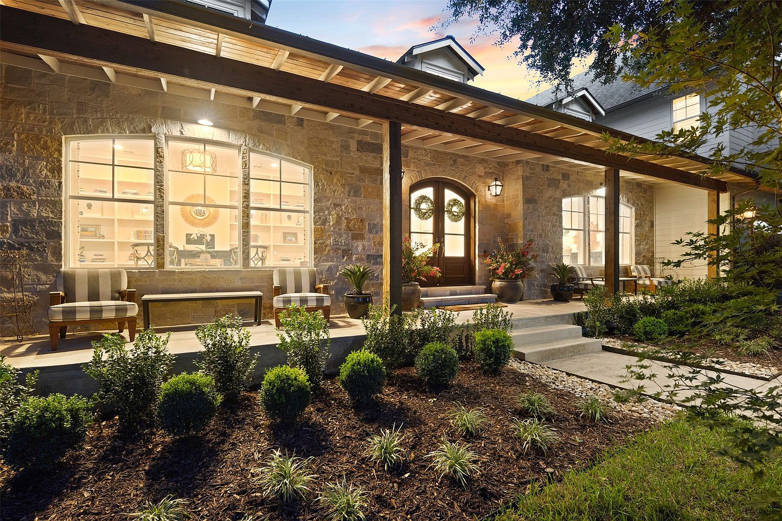 The image showcases the front exterior of a stone-clad house with a welcoming porch. The porch features wooden beams, comfortable seating, and well-manicured landscaping in the front yard. The overall impression is one of warmth, charm, and curb appeal, making it an attractive property.