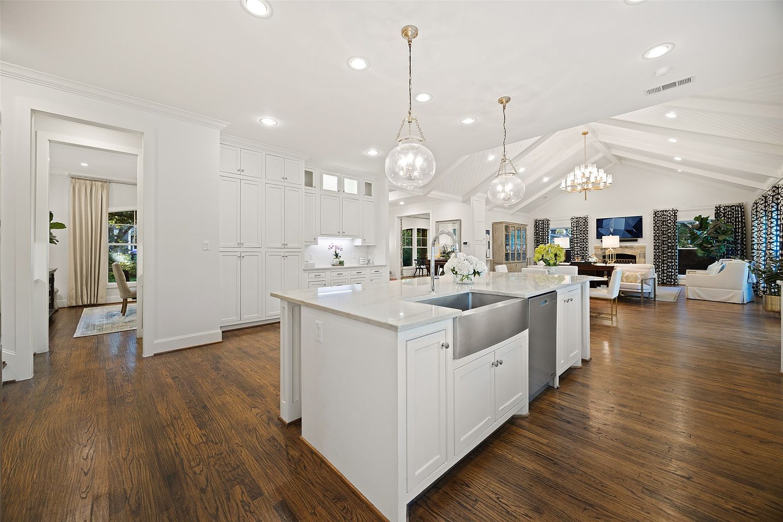 This is a bright and spacious kitchen featuring a large central island with a stainless steel farmhouse sink and white cabinetry. The kitchen opens into a living area with a vaulted ceiling and a fireplace, creating an open-concept living space. Hardwood floors run throughout, adding warmth and elegance to the home.