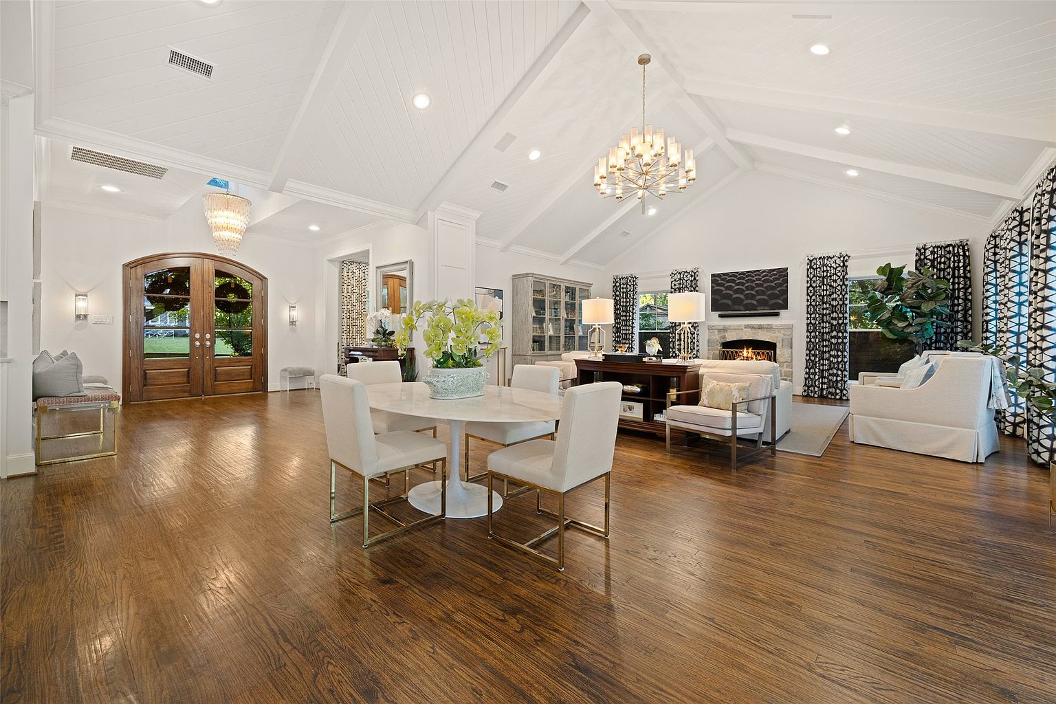 This is an interior shot of a spacious living room featuring hardwood floors, a vaulted ceiling with recessed lighting, and a stone fireplace. The room is furnished with a dining table, chairs, sofas, and a decorative cabinet, creating a comfortable and elegant atmosphere. Large windows with patterned curtains provide natural light and a view of the outdoors.