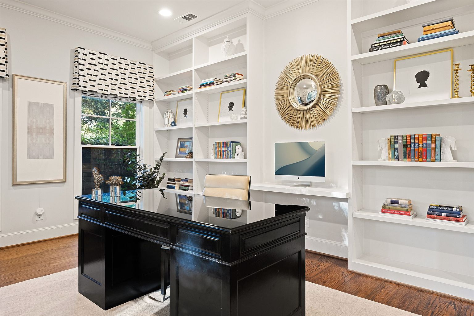 This is an interior shot of a home office featuring a large, black, glossy desk as the focal point. The room is well-lit, with natural light coming through a window with a patterned shade. Built-in white bookshelves line the wall, displaying books and decorative items, while a gold sunburst mirror and a modern computer add to the room's sophisticated and functional design.