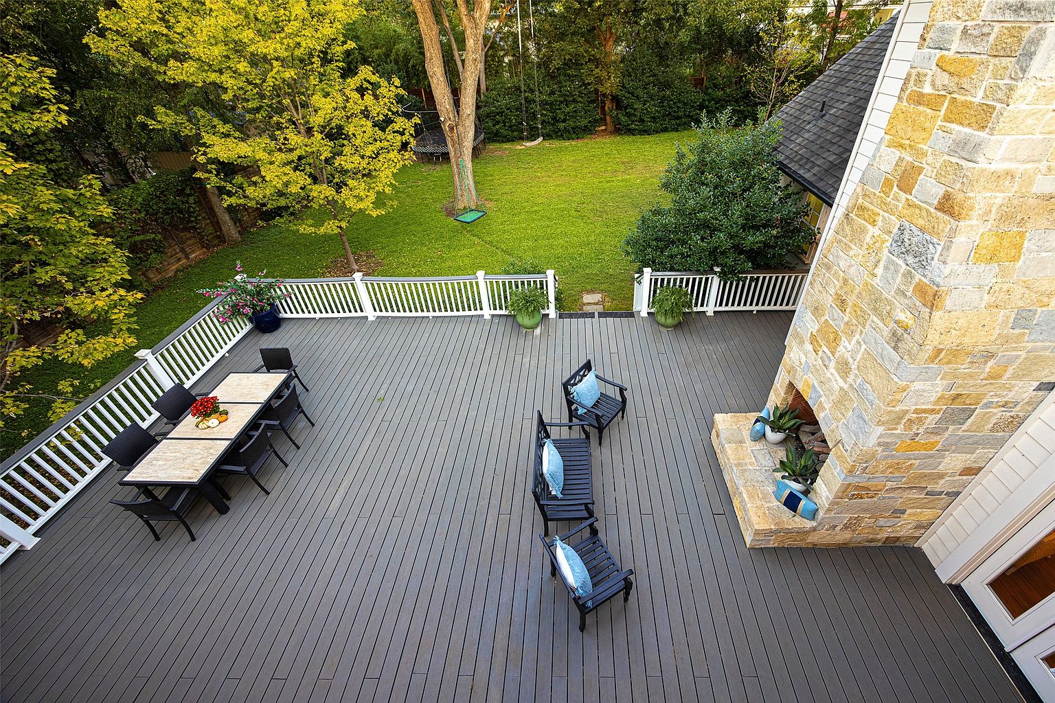 This is an aerial view of a spacious outdoor deck area, featuring a dining table with chairs, lounge seating with blue pillows, and a stone fireplace. The deck is surrounded by a white railing, offering views of a well-maintained lawn and mature trees. The overall impression is one of a luxurious and inviting outdoor living space.