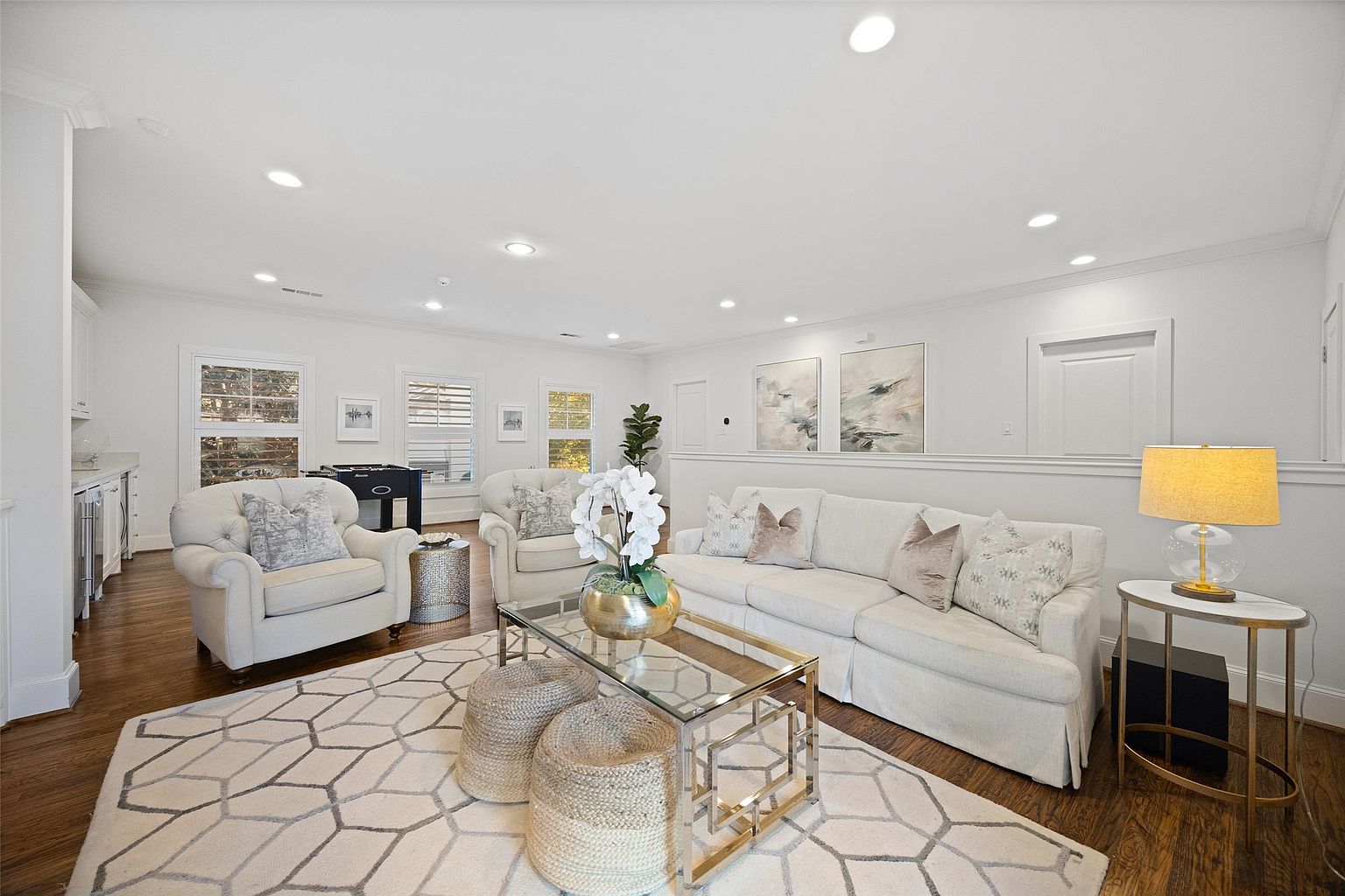 This is a bright and inviting living room featuring a neutral color palette with white walls and light-colored furniture. The room is well-lit with recessed lighting and natural light from the windows. Key features include a comfortable sofa, armchairs, a glass-topped coffee table, and a decorative area rug, creating a cozy and stylish space for relaxation and entertainment.