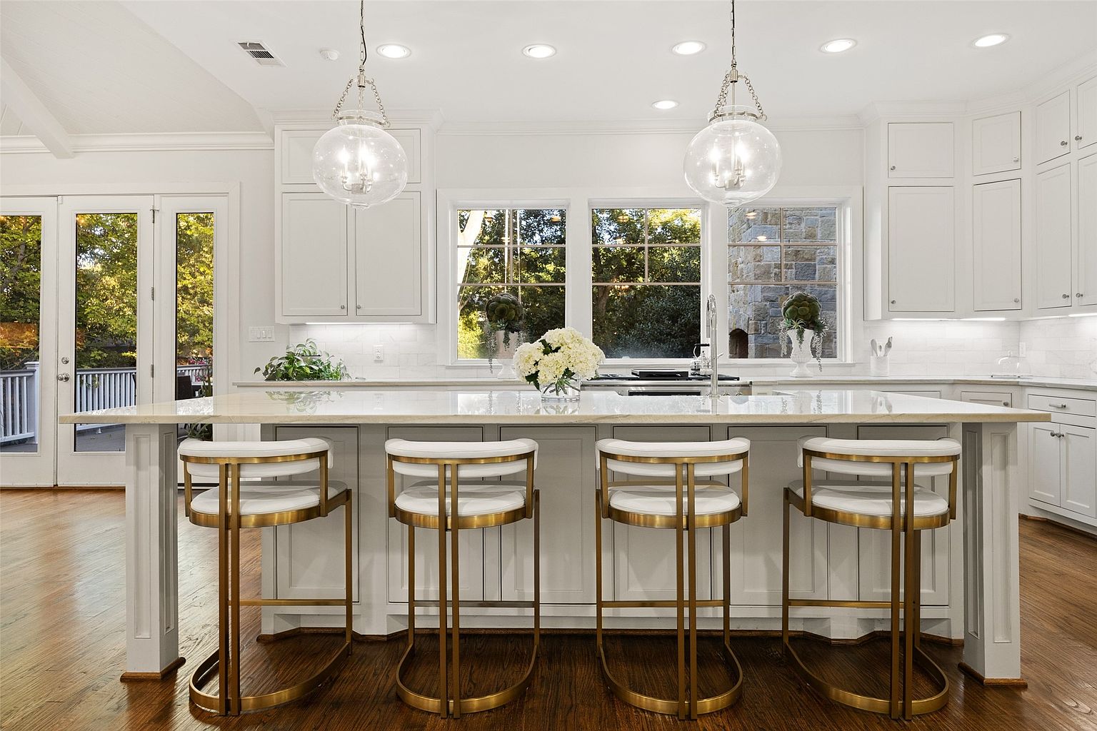This is a bright and airy kitchen featuring a large island with seating for four, illuminated by pendant lighting. The kitchen boasts white cabinetry, light countertops, and stainless steel appliances, creating a clean and modern aesthetic. Large windows provide natural light and a view of the outdoors, enhancing the inviting atmosphere.