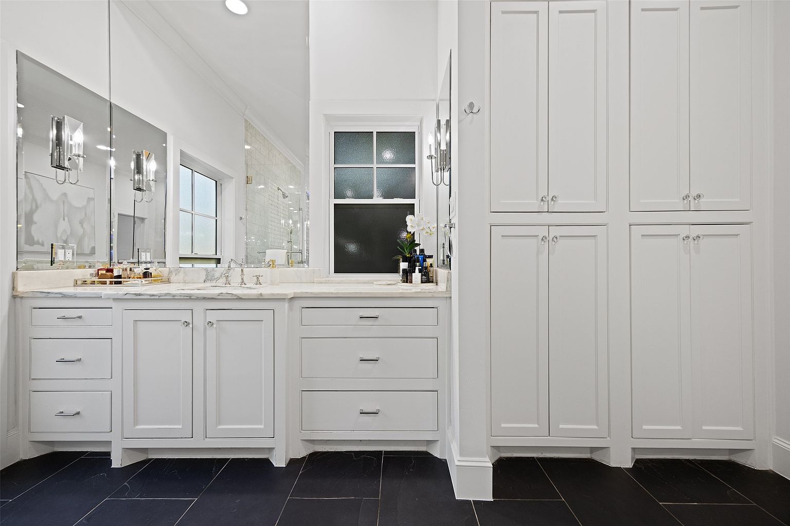 This is a well-lit primary bathroom featuring white cabinetry with sleek hardware and a marble countertop. A large mirror reflects the light, enhancing the spacious feel, while the dark flooring provides a striking contrast. The built-in storage cabinets offer ample space, contributing to the bathroom's organized and luxurious aesthetic.