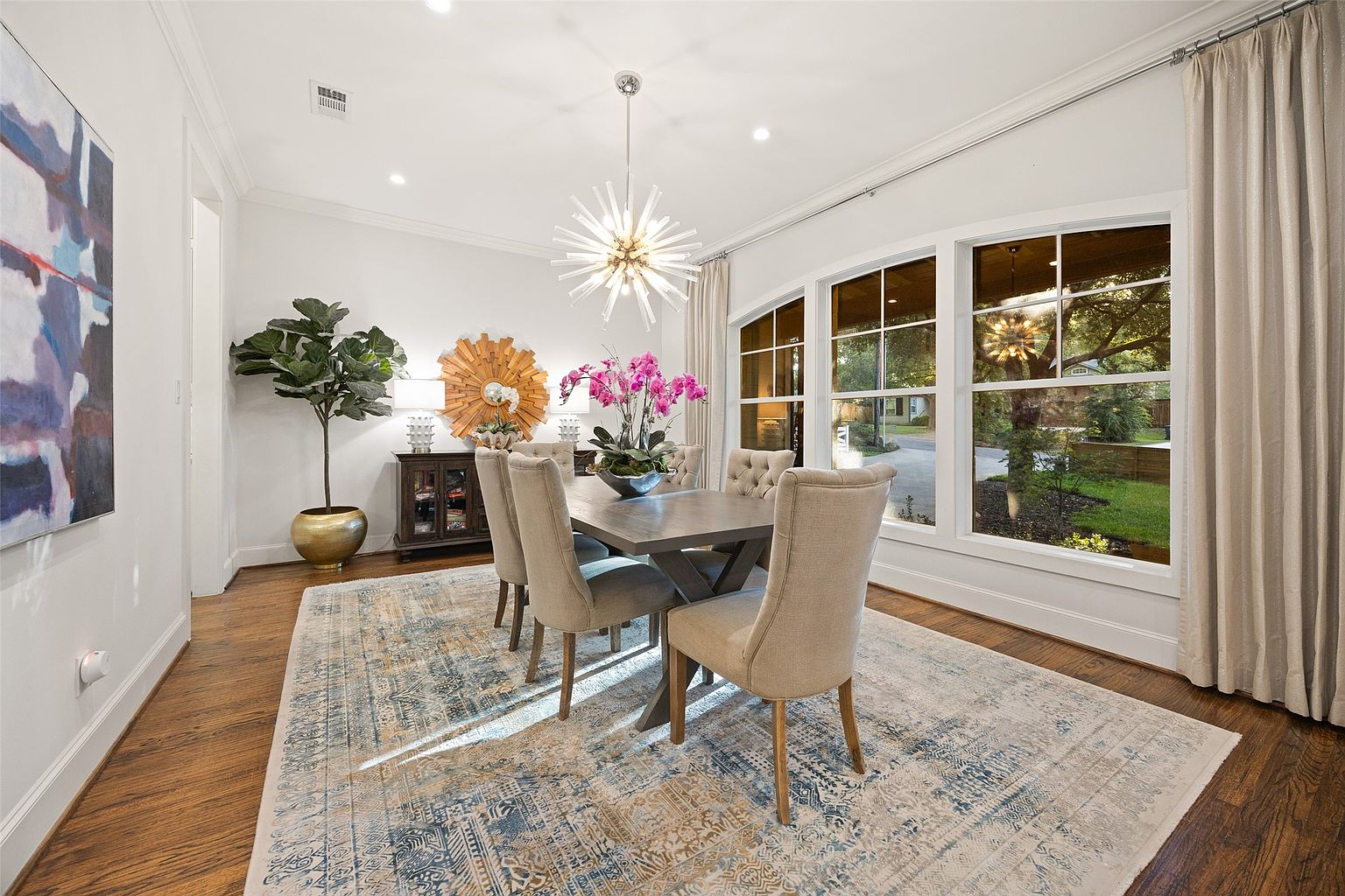 This is an interior shot of a dining room featuring a wooden dining table with upholstered chairs, a modern chandelier, and a large window offering natural light. The room is decorated with a large area rug, a decorative mirror, and a potted plant, creating a warm and inviting atmosphere. The walls are painted in a neutral color, and the hardwood floors add to the room's elegance.