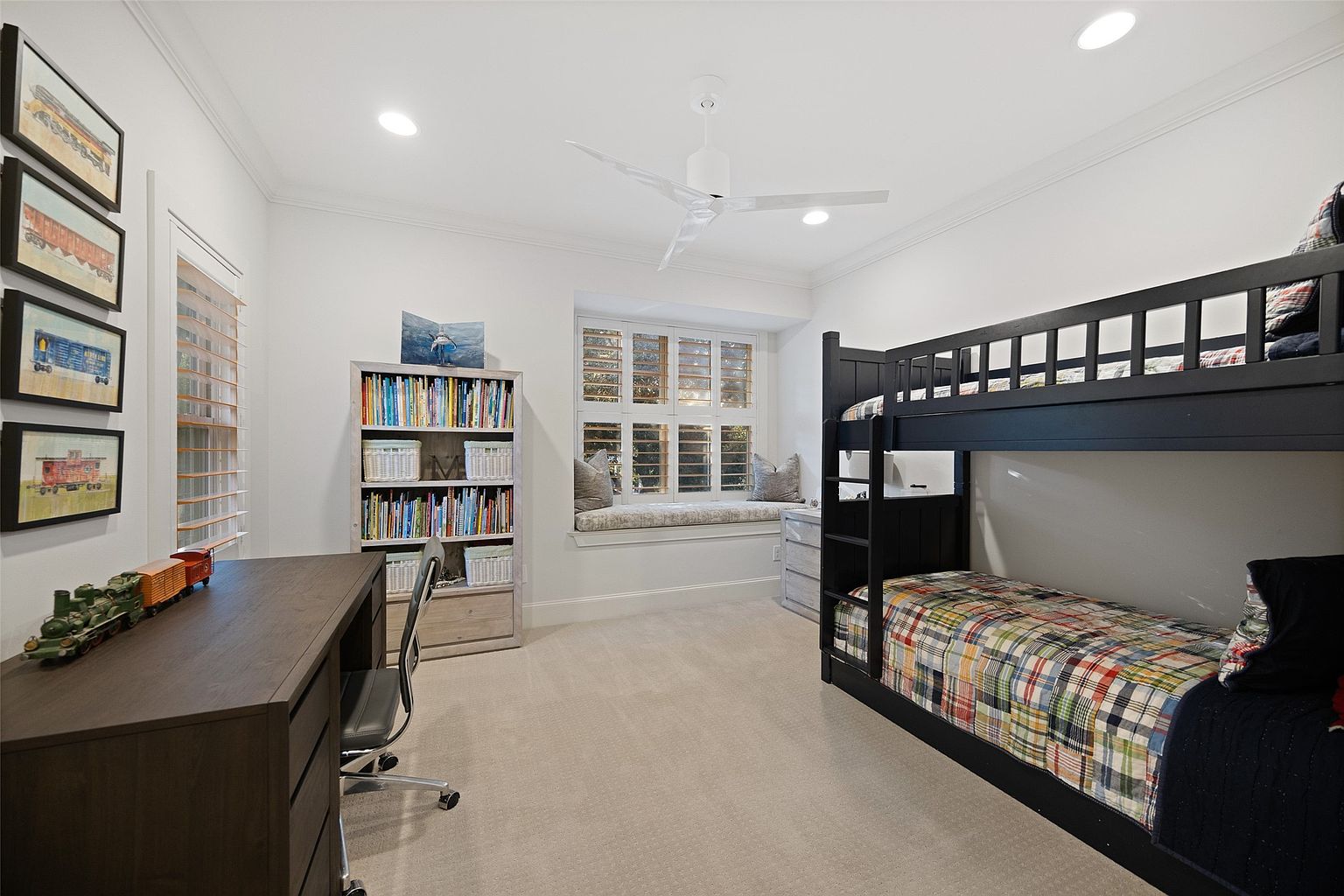 This is a well-lit bedroom featuring a dark wood bunk bed with a colorful plaid quilt on the bottom bunk. A wooden desk with a rolling chair sits to the left, accompanied by a bookshelf filled with books and baskets. A window seat with patterned cushions is nestled between the bookshelf and the bunk bed, creating a cozy and inviting space.