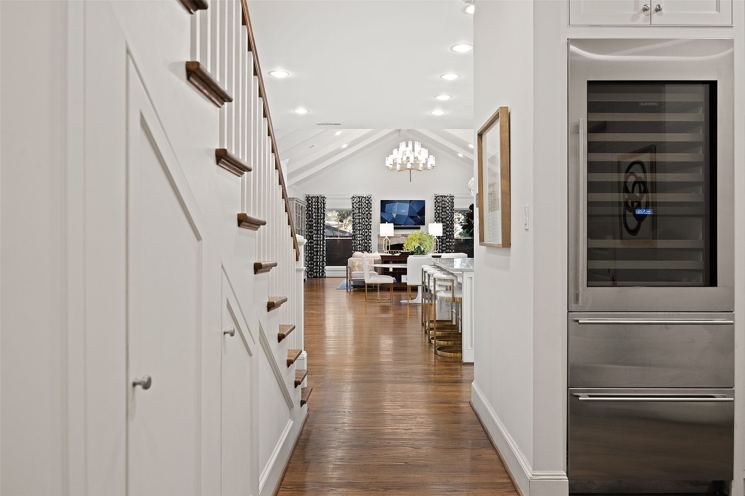 This interior shot showcases a hallway leading into a bright living space. The hallway features hardwood floors, white walls, and a staircase with wooden accents. A built-in wine fridge is visible on the right, adding a touch of luxury, while the living room beyond boasts a chandelier and ample natural light, creating an inviting atmosphere.