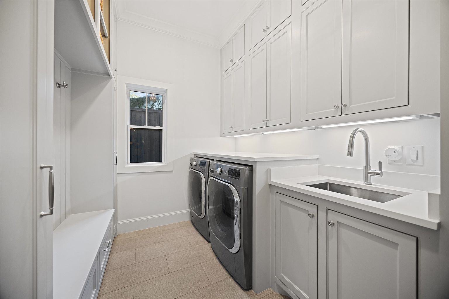 This is a well-lit laundry room featuring a side-by-side washer and dryer set, a stainless steel sink with a modern faucet, and ample white cabinetry above and below the countertop. A window provides natural light, and the room includes a built-in bench and storage area. The overall impression is clean, organized, and functional.